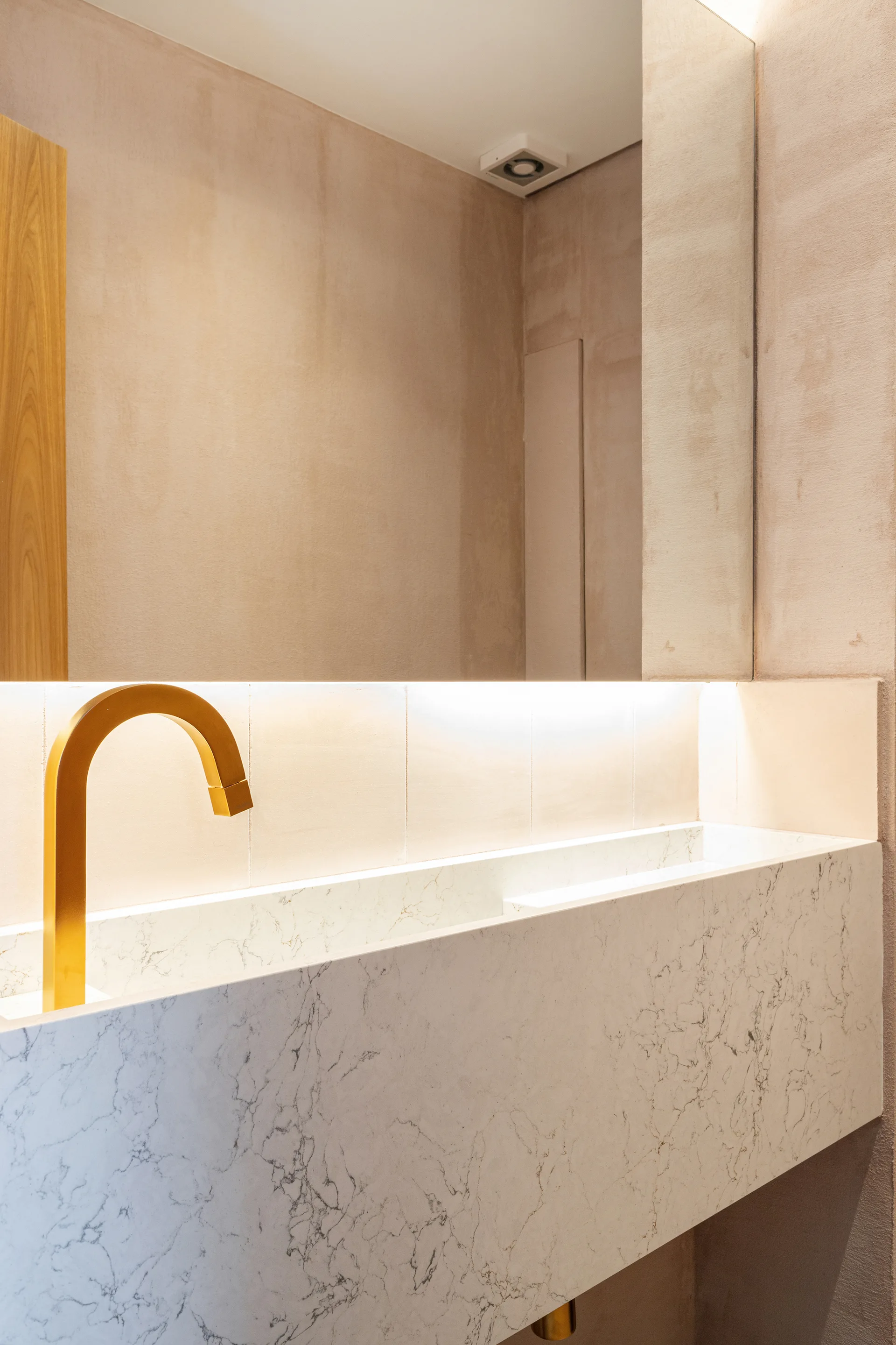 A modern bathroom features a veined white marble trough sink with an arched gold faucet beneath a large wall mirror with integrated glowing light.