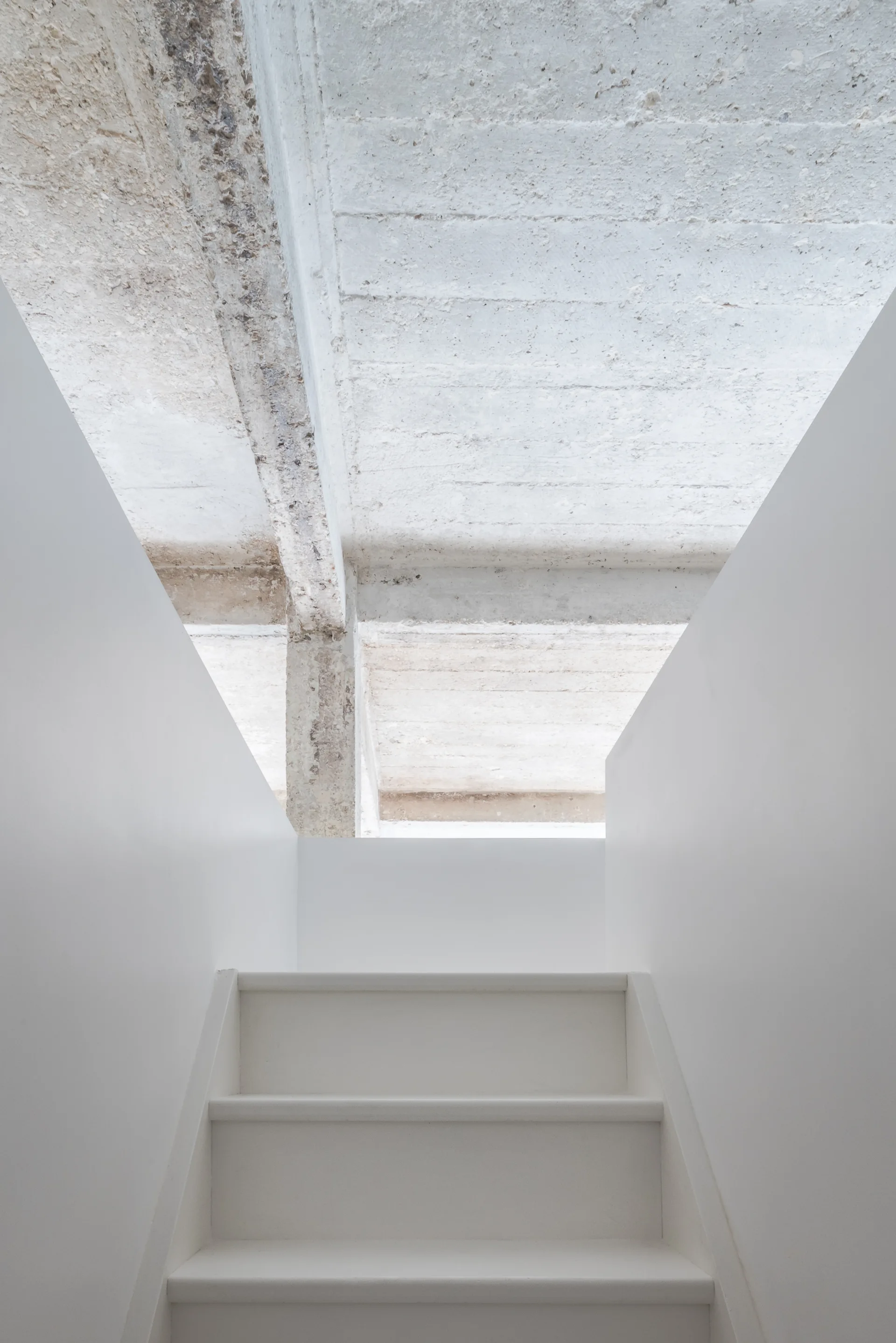 A minimalist white staircase with smooth white walls ascends toward an unfinished, rough gray concrete ceiling with structural beams.