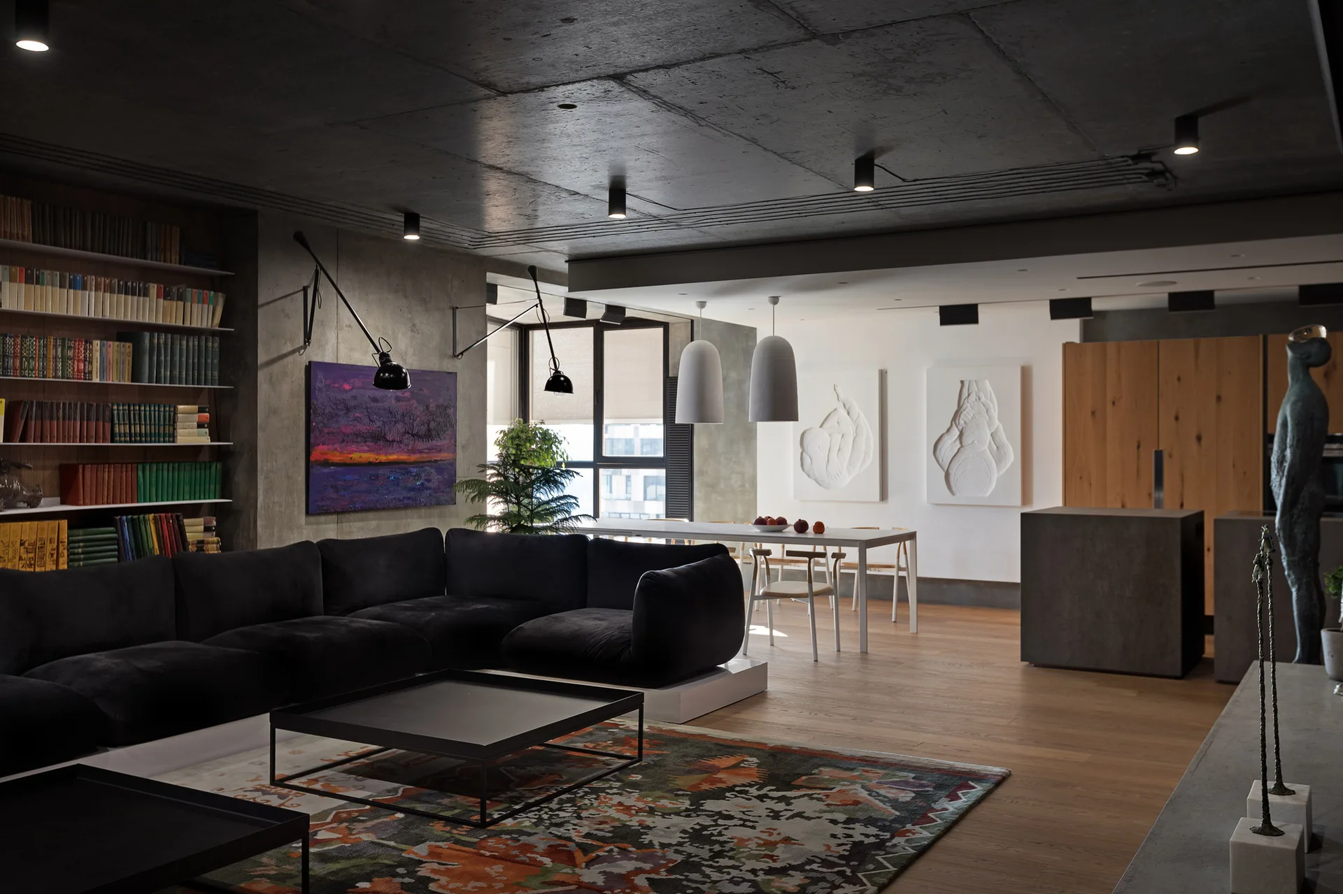 A contemporary open-plan living area features a large black sectional sofa, a patterned rug, an exposed concrete ceiling, and a wooden built-in bookshelf.
