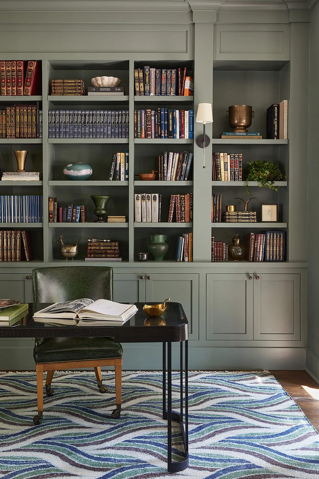 A home office features sage green built-in bookshelves filled with books, a dark wood desk with a leather chair, and a wavy patterned blue and green rug.