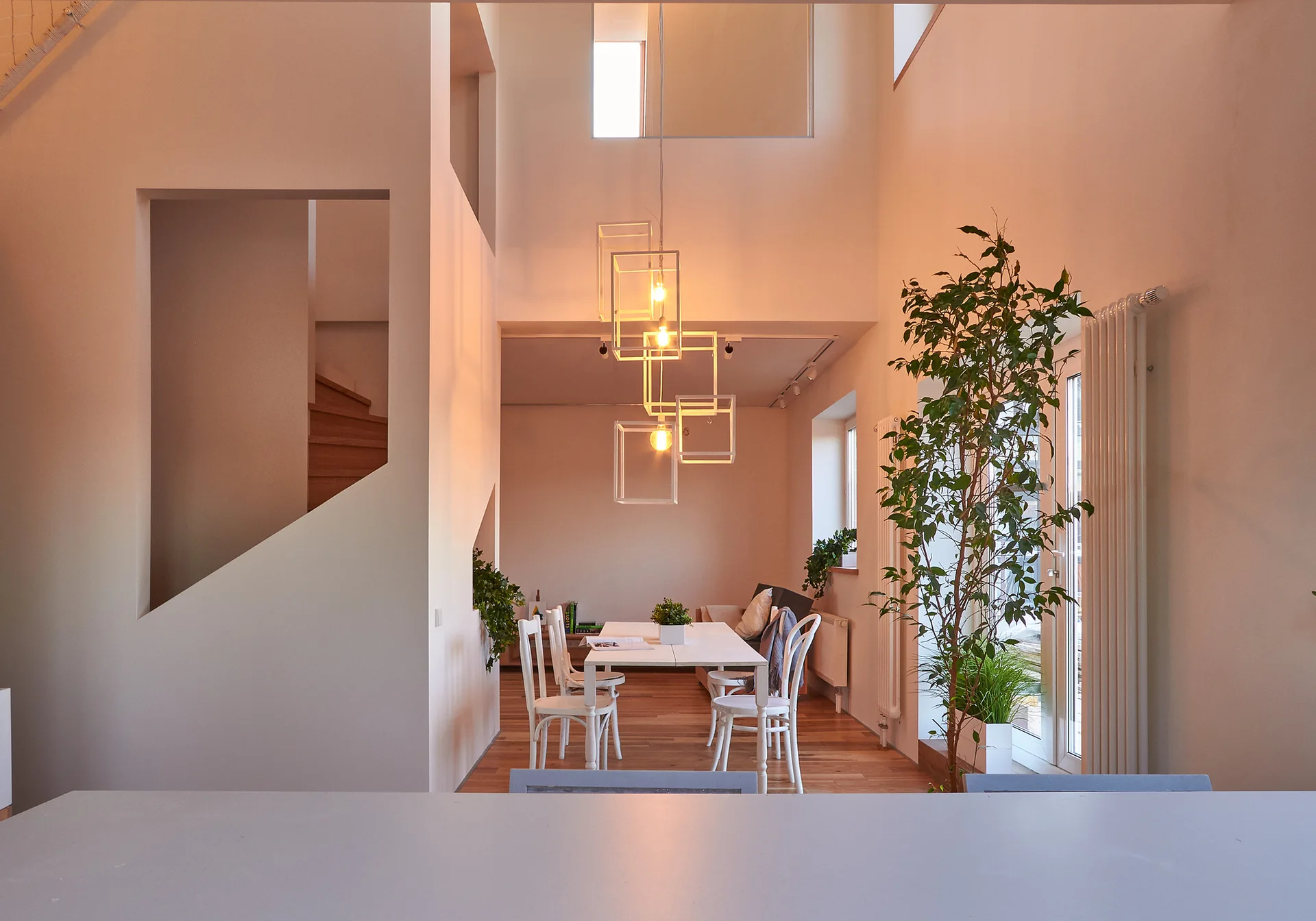 A white minimalist dining room features a dining table with chairs, wireframe pendant lights, and a wooden staircase visible through a wall opening.