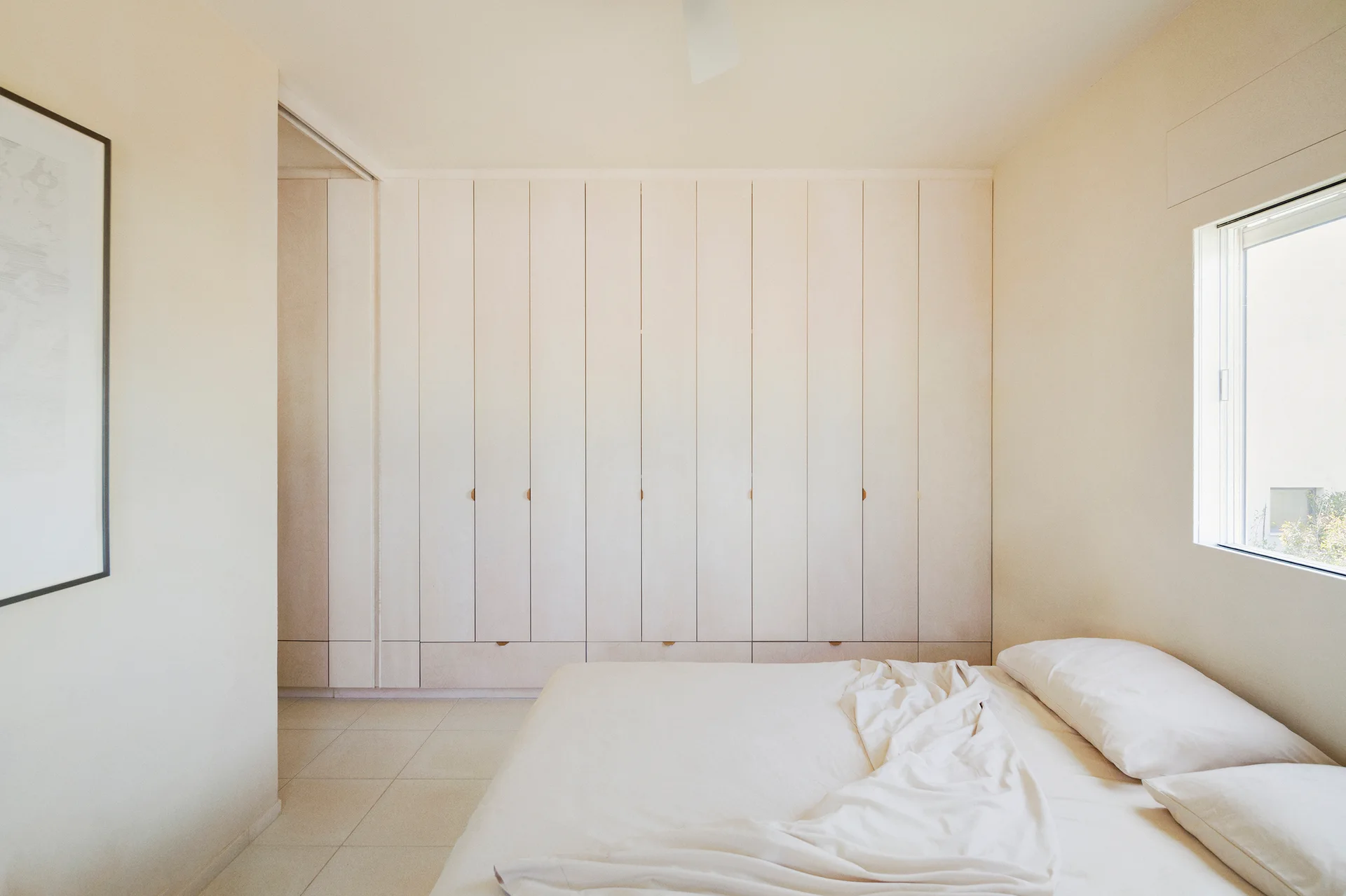 A minimalist cream bedroom features a bed with off-white linens, a large-format tile floor, and a full-wall built-in wardrobe with flat panels.