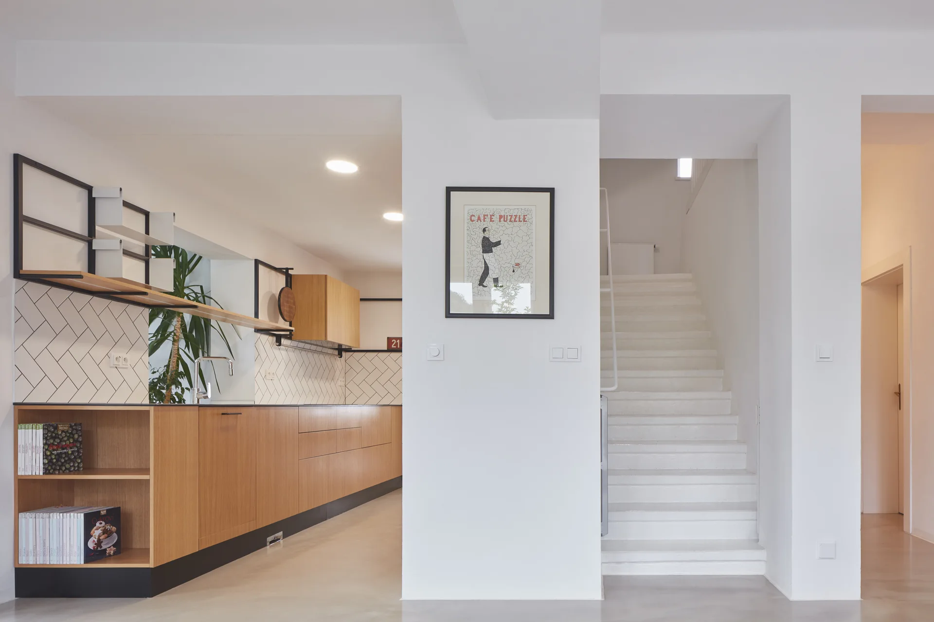 A minimalist kitchen featuring wood base cabinets, a white herringbone tile backsplash, and an adjacent white staircase.