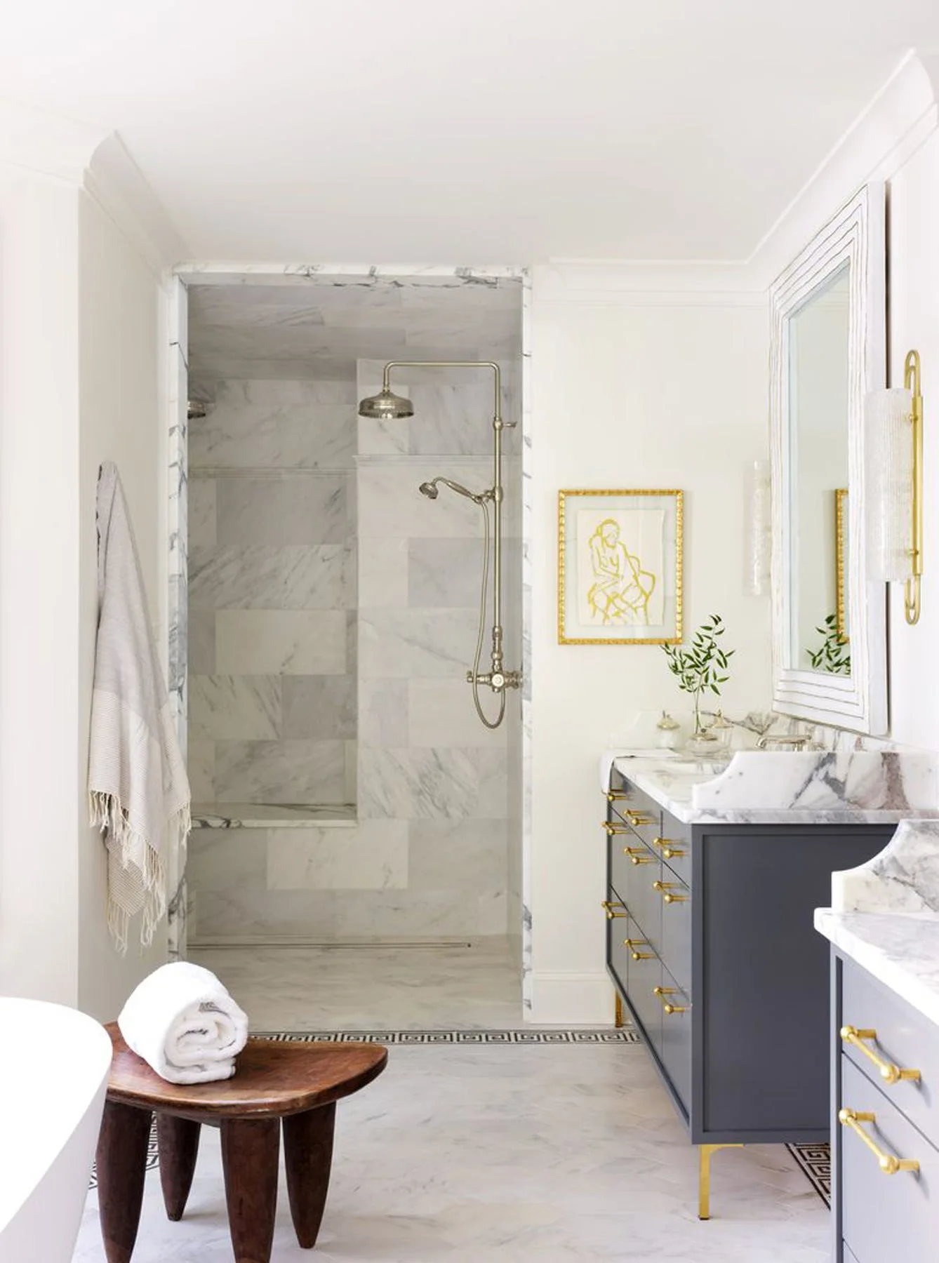 A bright bathroom featuring a dark gray vanity with gold hardware, a marble-tiled walk-in shower, and a dark wood stool on a herringbone floor.