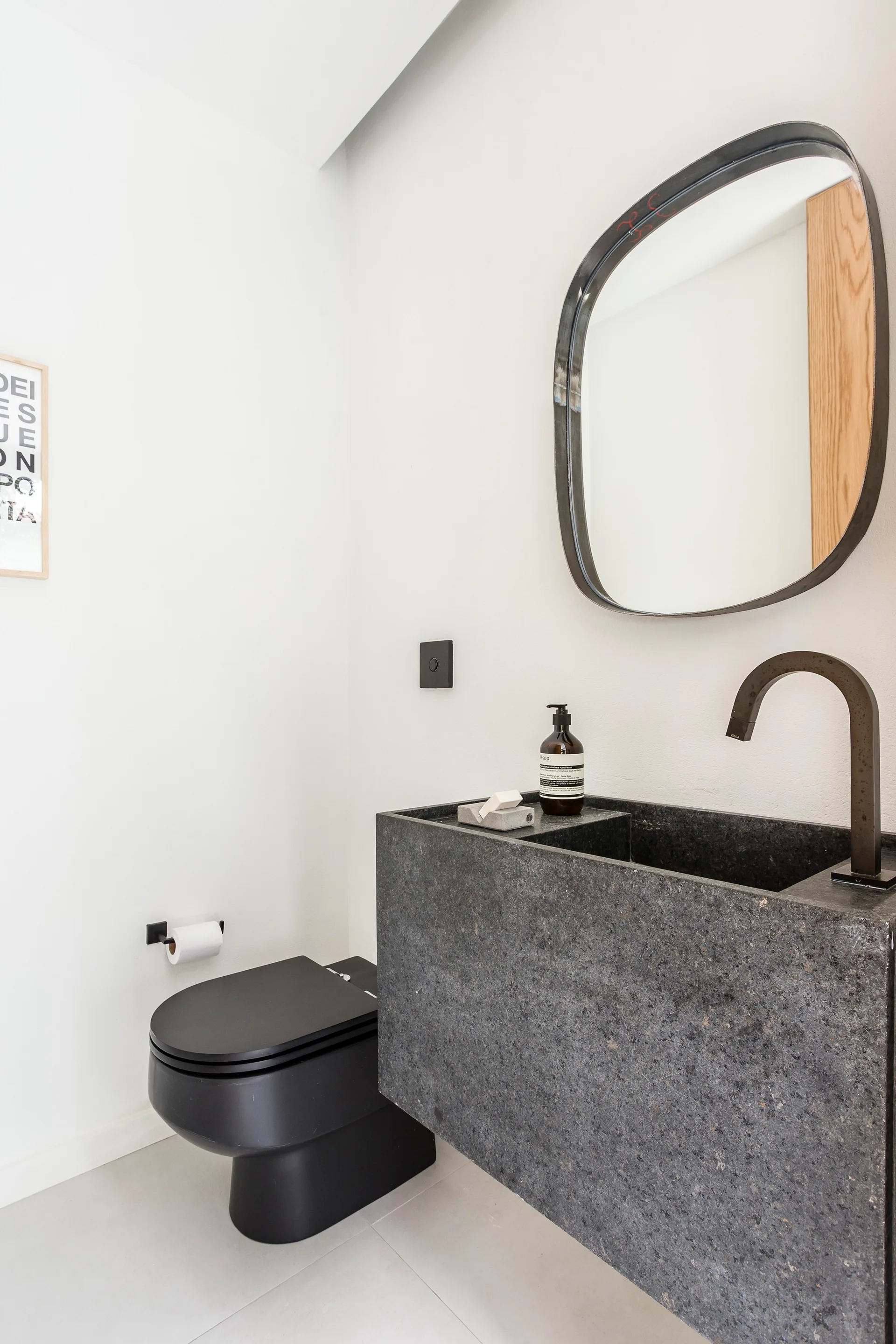 A minimalist powder room with a wall-mounted dark stone vanity, a matte black toilet, and an organically shaped mirror on a white wall.