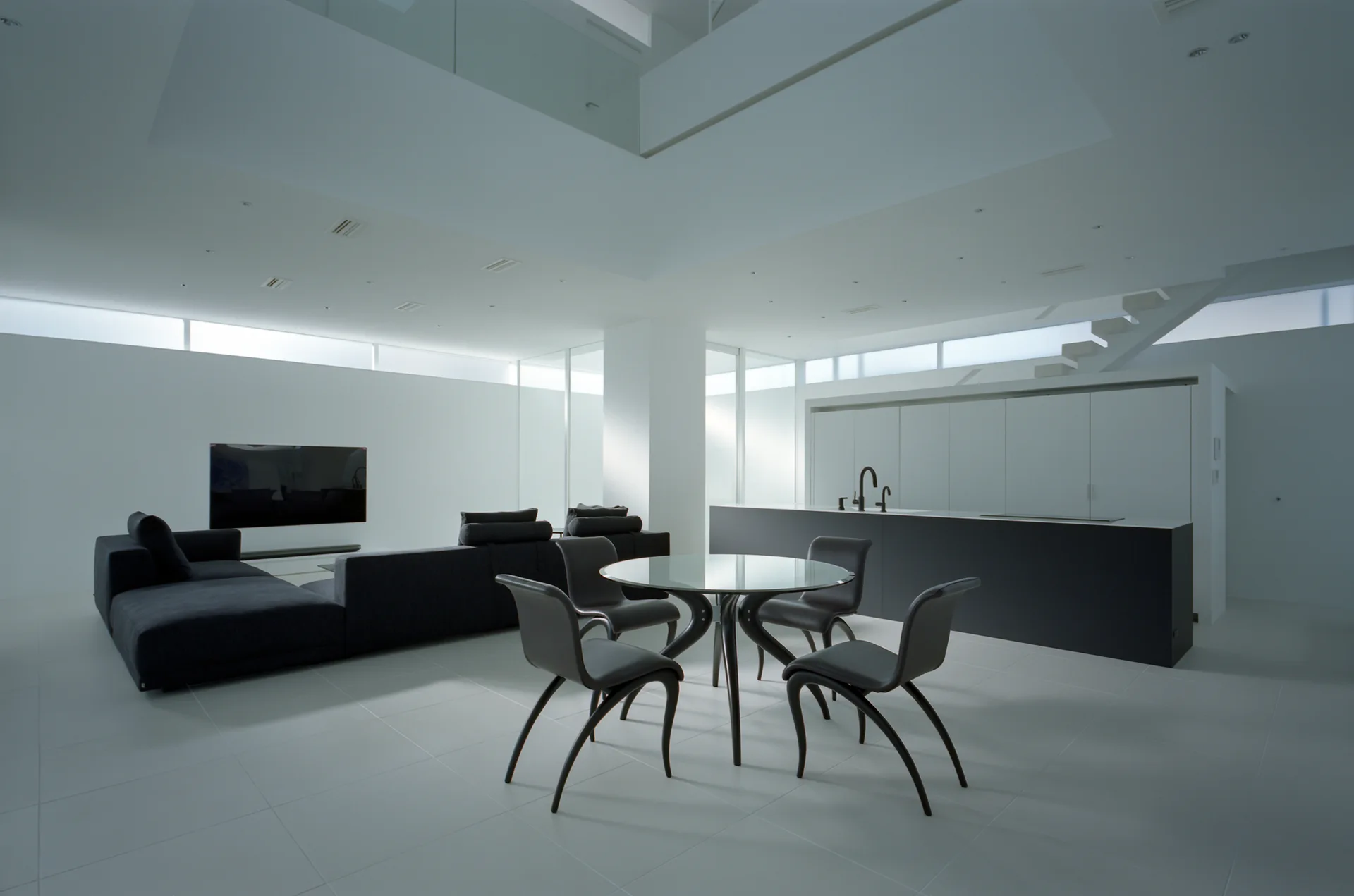 A minimalist double-height white room features a black sectional sofa, a round glass dining table with curved black chairs, and a black kitchen island.