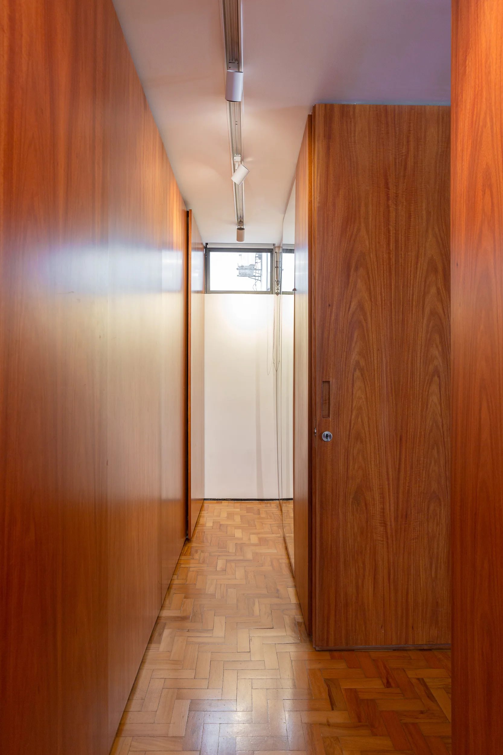 A narrow hallway featuring floor-to-ceiling wood paneled walls, a herringbone parquet floor, and a single track light leading to a small window.