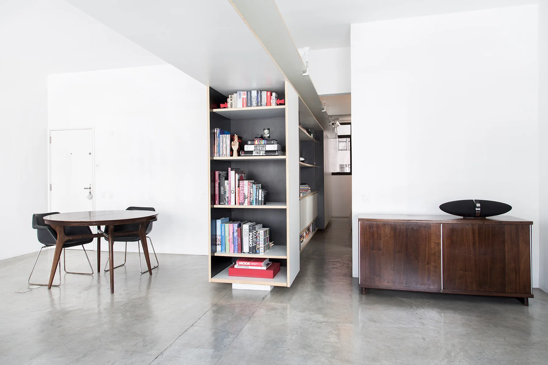 A minimalist apartment interior featuring a central dark bookshelf room divider, a wooden dining table, and polished concrete floors.