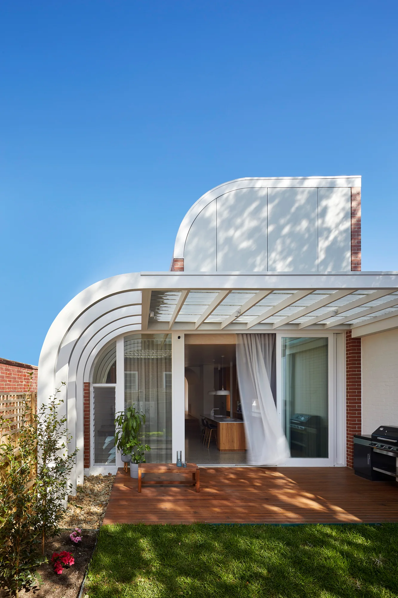 A modern backyard patio features a wooden deck, a curved white architectural pergola with a ribbed roof, and sliding glass doors.