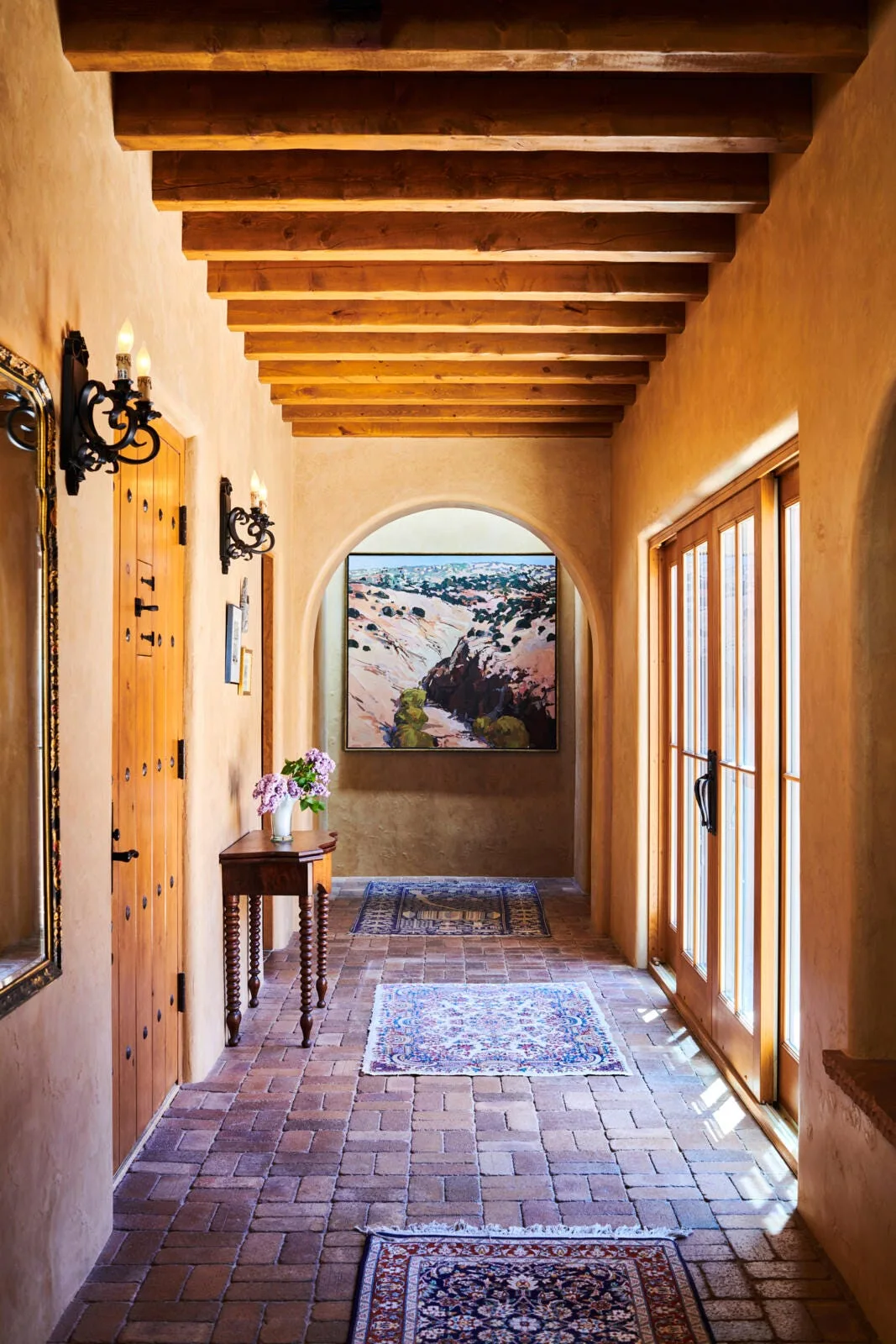 A rustic hallway featuring textured terracotta plaster walls, a brick floor lined with patterned rugs, exposed wooden ceiling beams, and an arched doorway.