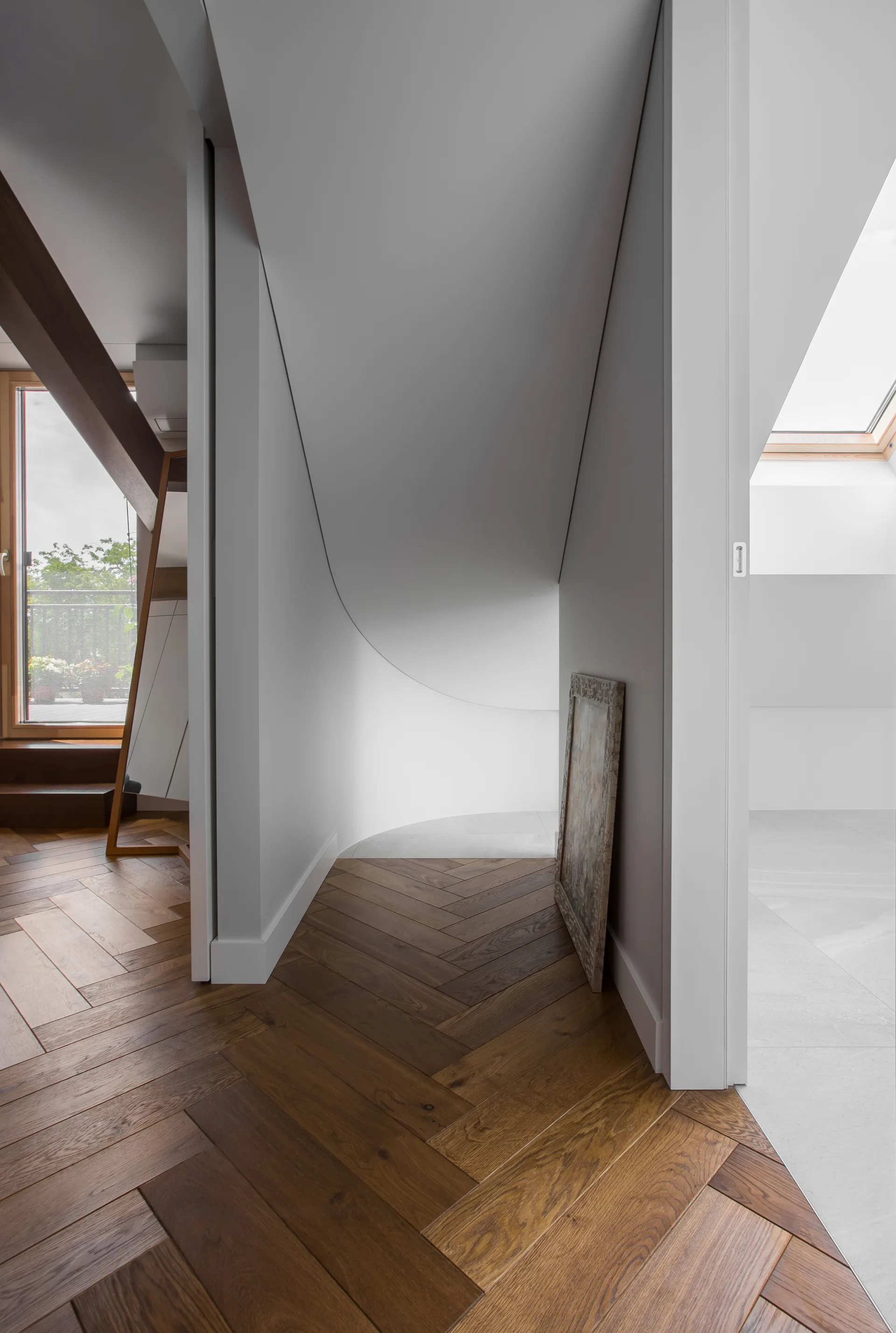 A modern attic hallway features medium brown herringbone hardwood floors, a curved white partition wall, and a slanted ceiling with a skylight.