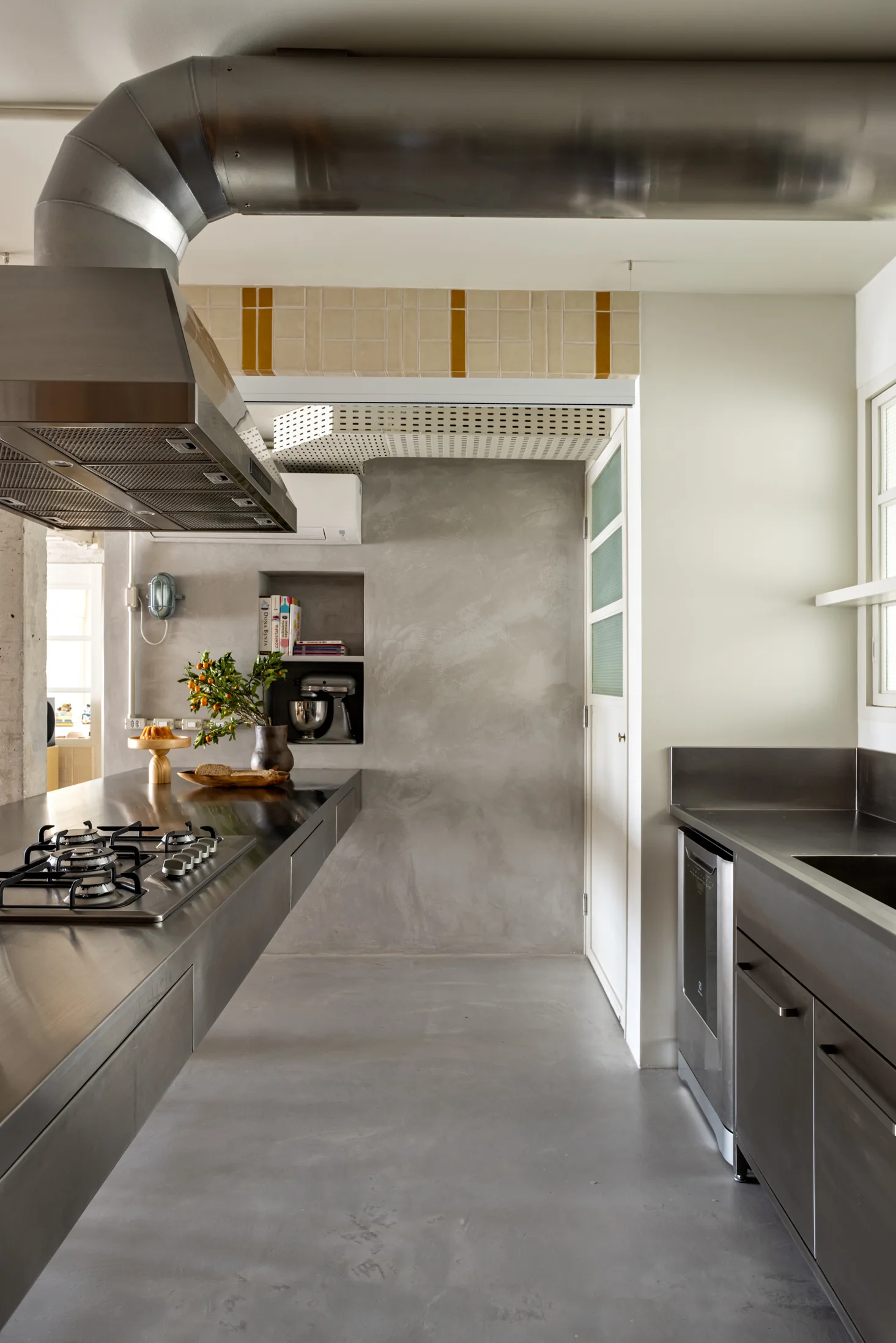 An industrial kitchen features a stainless steel island, concrete walls, a recessed wall niche, and exposed metal ventilation ducts.