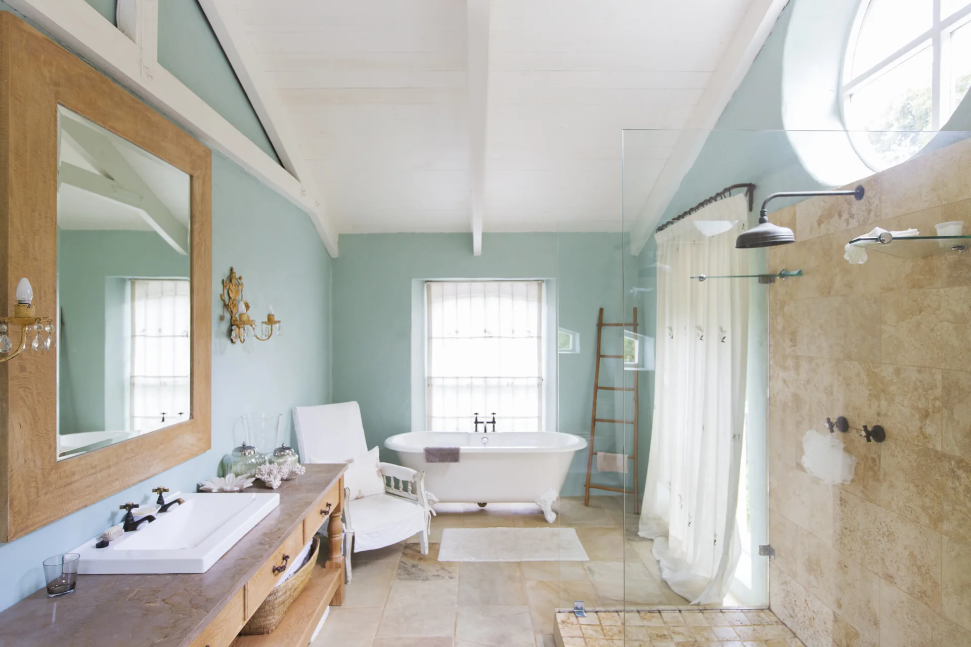 A bright cottage bathroom features a white clawfoot bathtub, pale blue walls, a wooden vanity, and a glass-enclosed shower with stone tile.