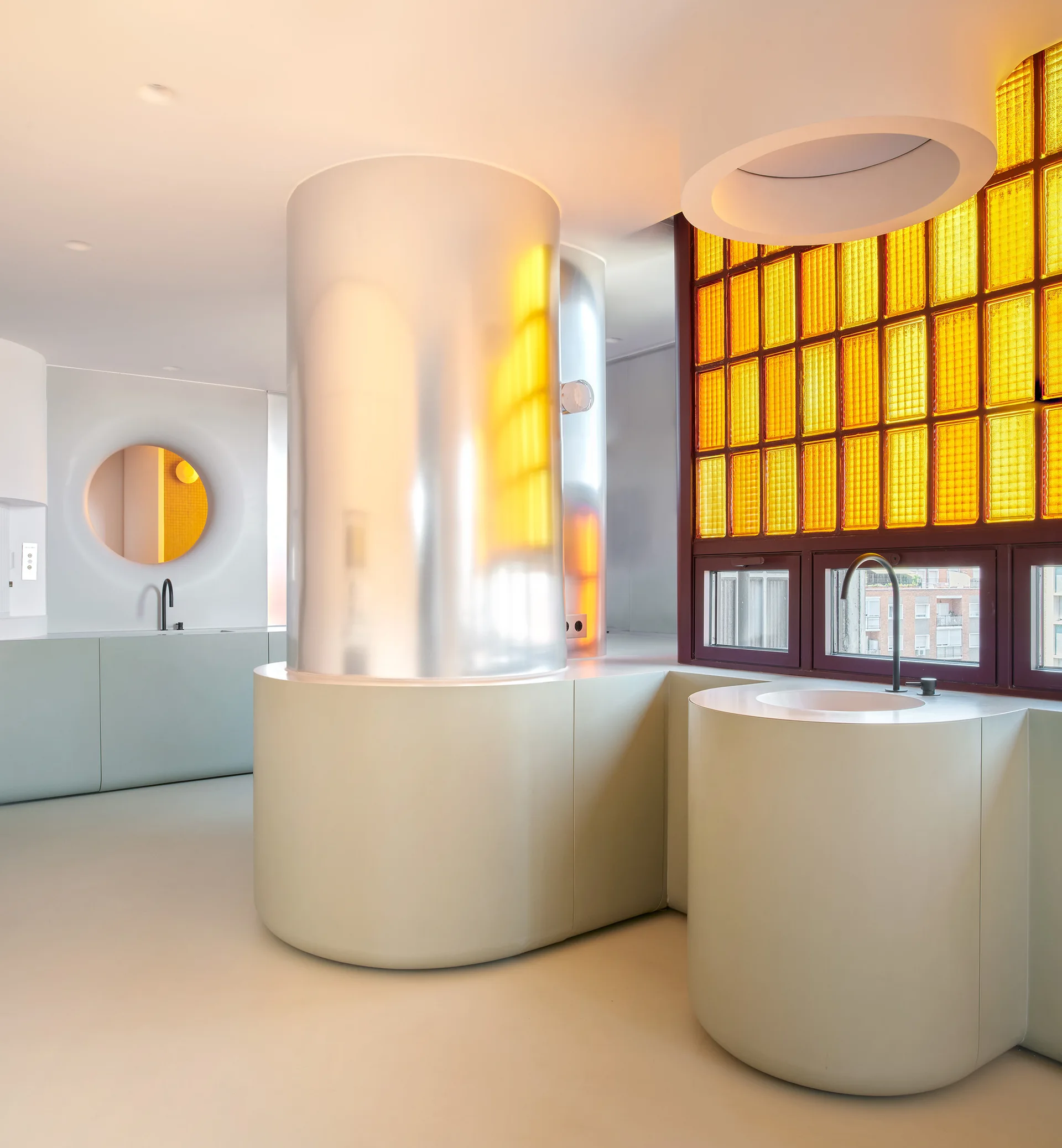 A minimalist kitchen features curved pale green cabinetry, a reflective silver column, and a grid window with bright yellow glass panes.