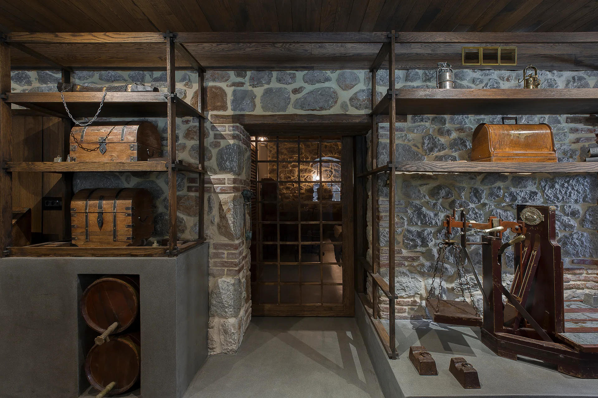 A rustic cellar room features exposed stone walls, dark wood plank ceilings, open wooden shelving holding antique chests, and a vintage scale on the floor.