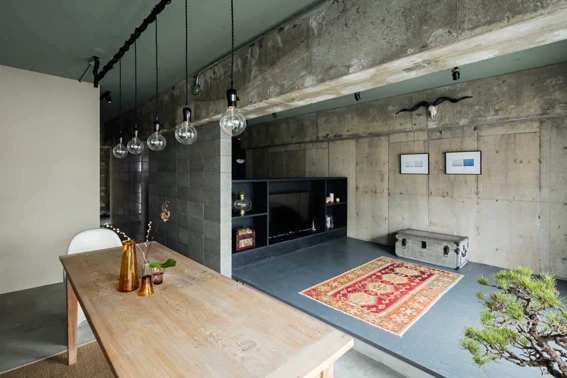 An industrial interior features raw concrete walls, a raised living area with a patterned rug, and a rustic wooden dining table with pendant lights.
