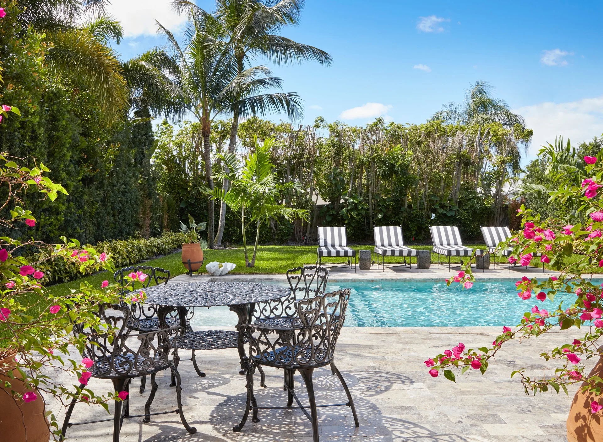 A sunny backyard pool area features a black wrought iron dining table set on a stone patio, with striped lounge chairs and tropical landscaping.
