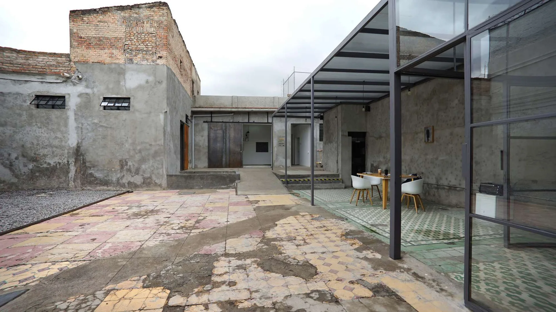 A weathered concrete courtyard features a metal-framed patio cover shading a wooden dining table with white chairs on distressed patterned tiles.