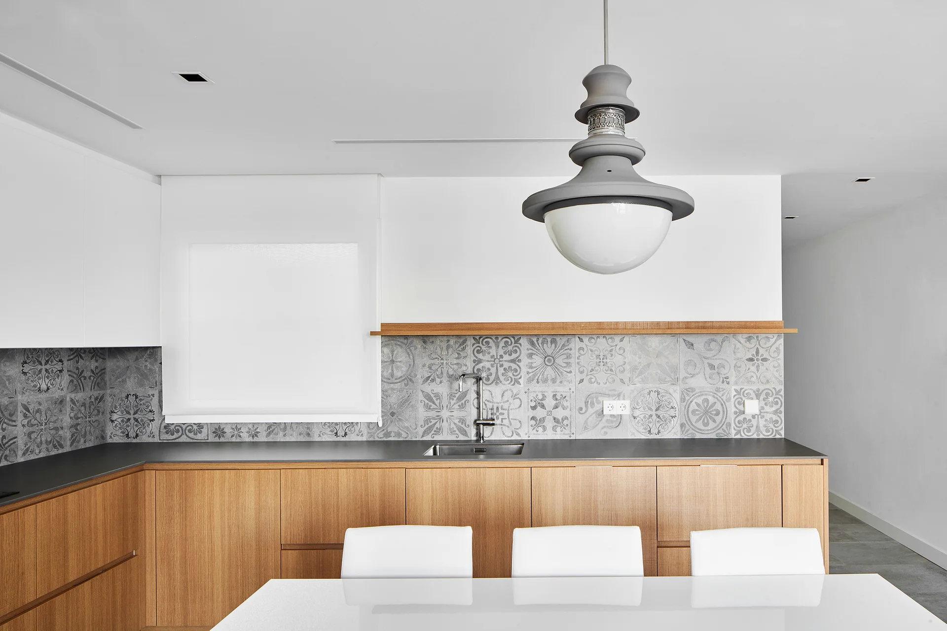 A modern kitchen features light wood cabinets, an intricate grey patterned tile backsplash, a dark countertop, and a large sculptural pendant light.
