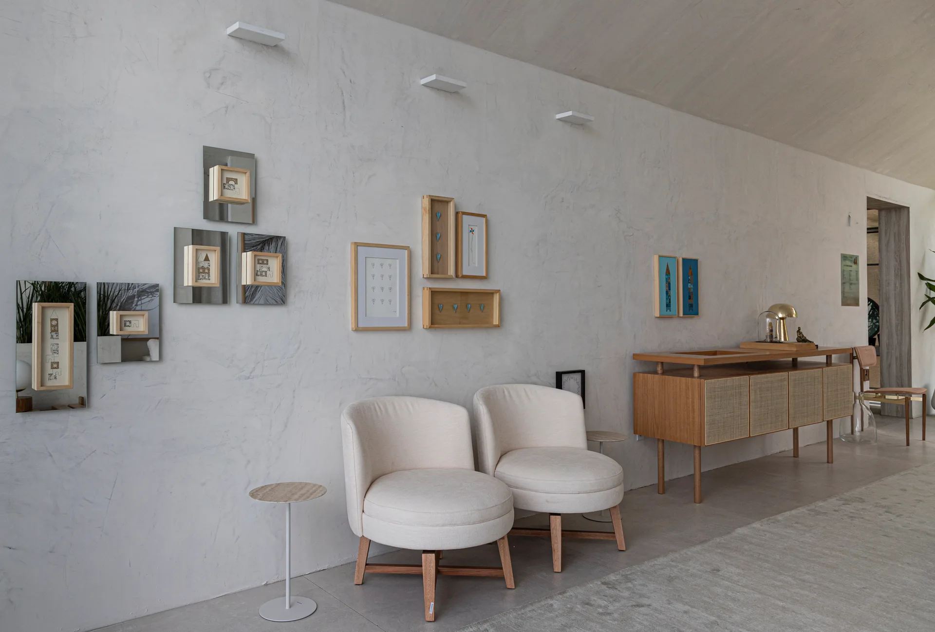 Two cream rounded armchairs and a wooden cane sideboard sit against a textured plaster wall decorated with a unique gallery of framed artwork.
