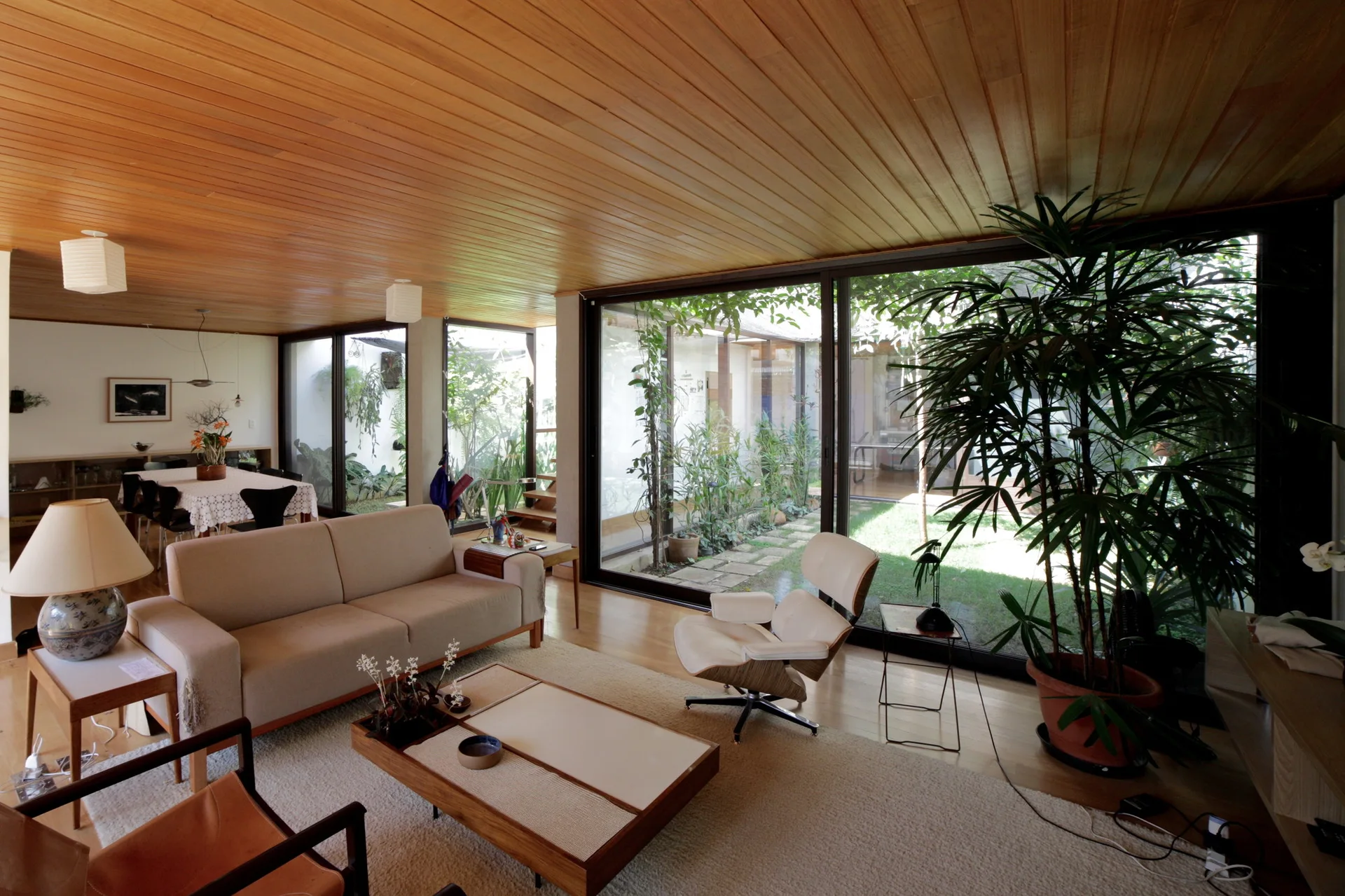 A mid-century modern living room features a warm wood-plank ceiling, a beige sofa, and large glass walls overlooking a green courtyard.