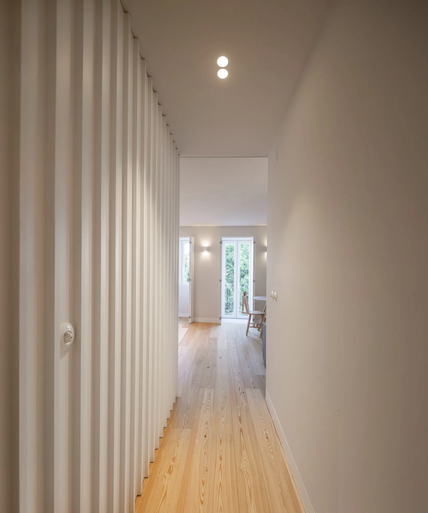 A long minimalist hallway with a white vertical slatted wall, light wood flooring, and recessed ceiling lights leading to a bright, sunlit room.