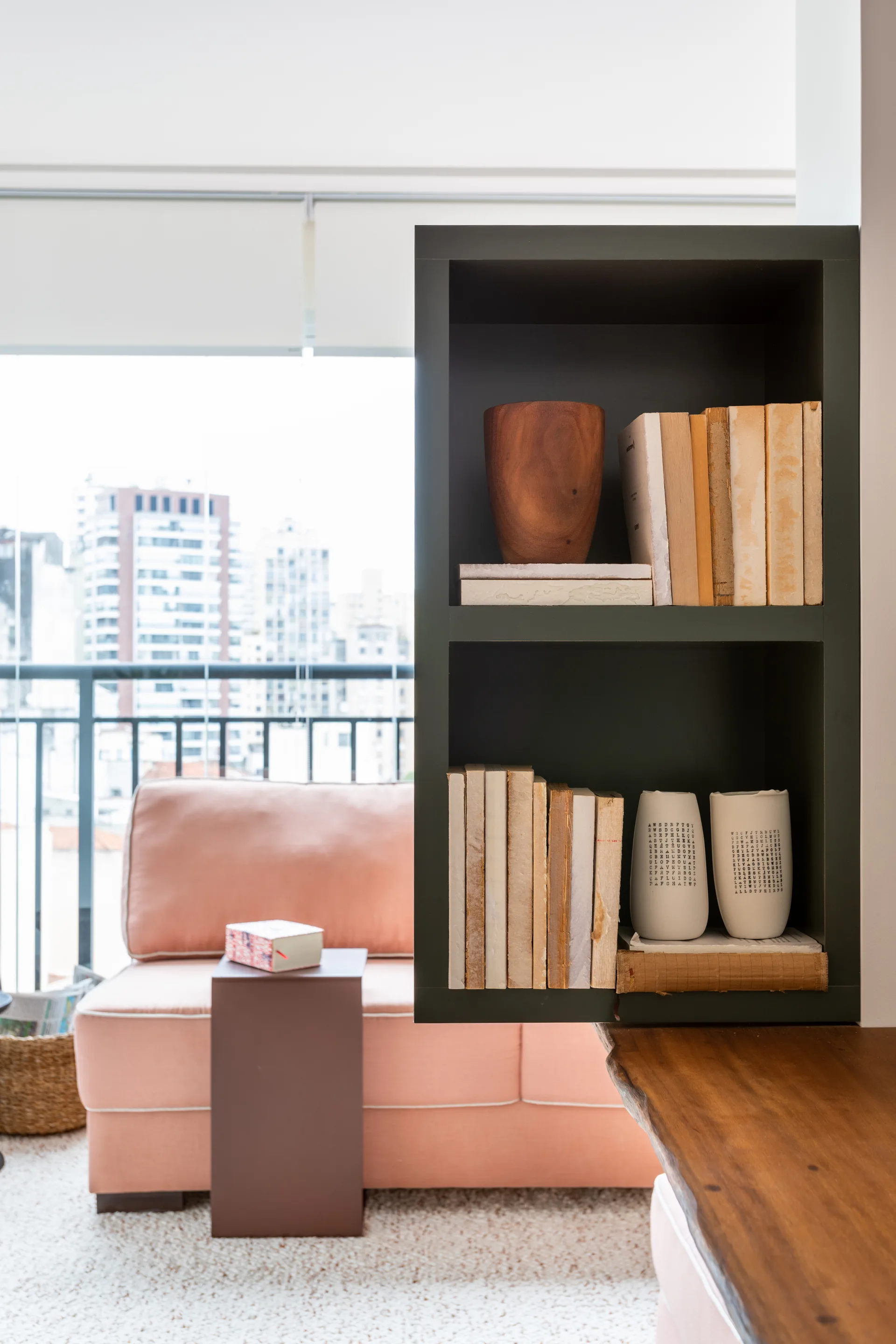 A dark green open bookshelf styled with books and ceramic vases stands next to a light pink sofa in a bright apartment living room.