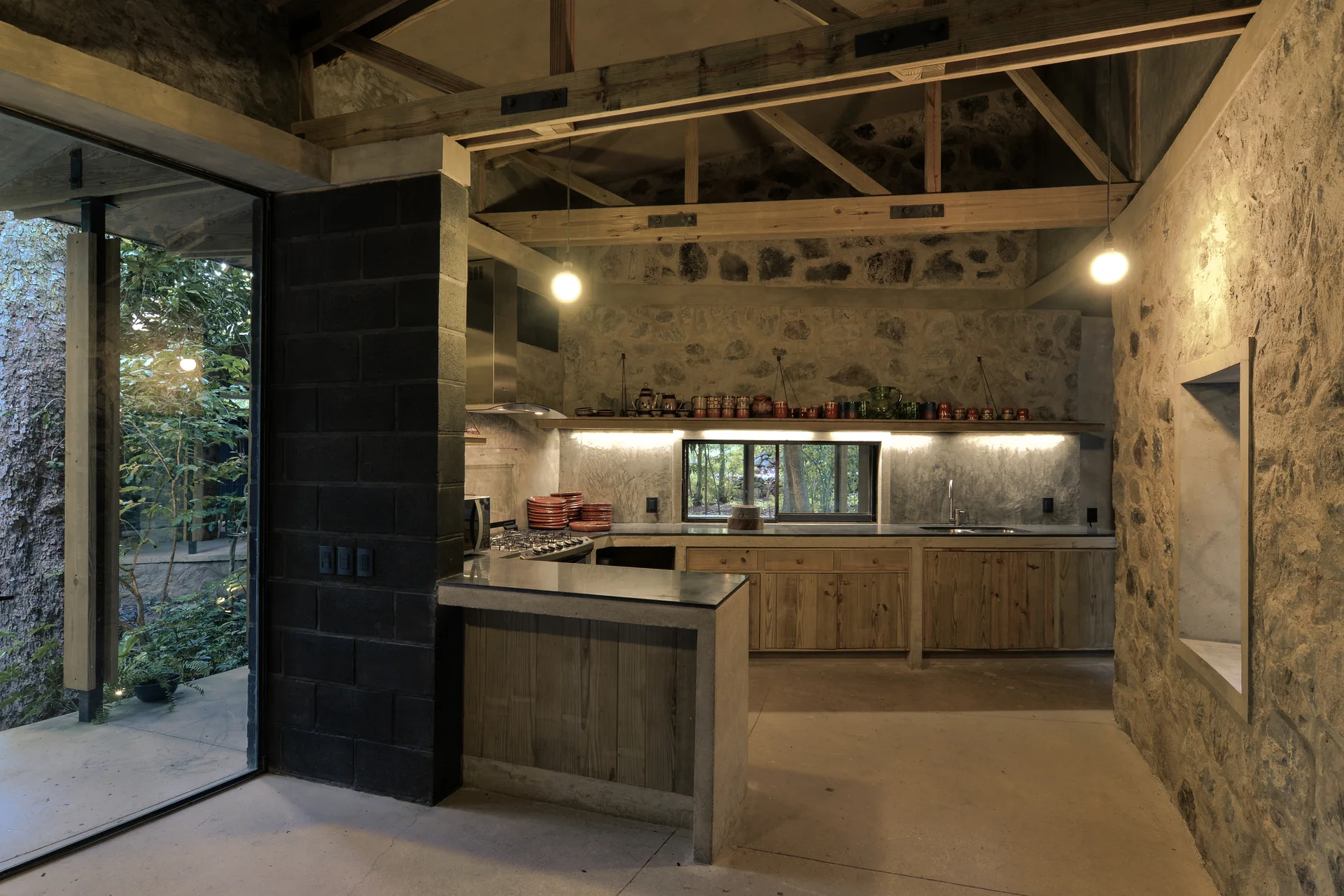 A rustic kitchen features natural stone masonry walls, exposed wooden ceiling trusses, concrete countertops, and simple wood cabinets.