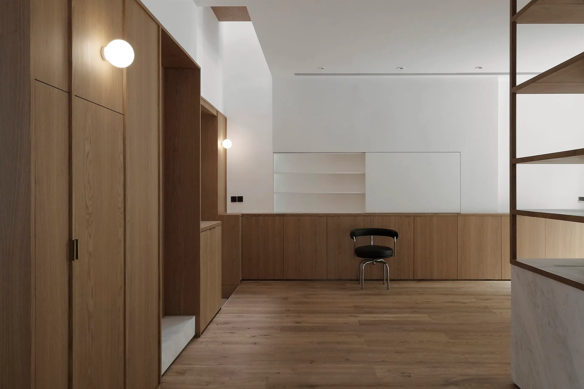 A minimalist living room features warm wood-toned built-in cabinetry, smooth wood flooring, white walls, spherical wall sconces, and a single black chair.