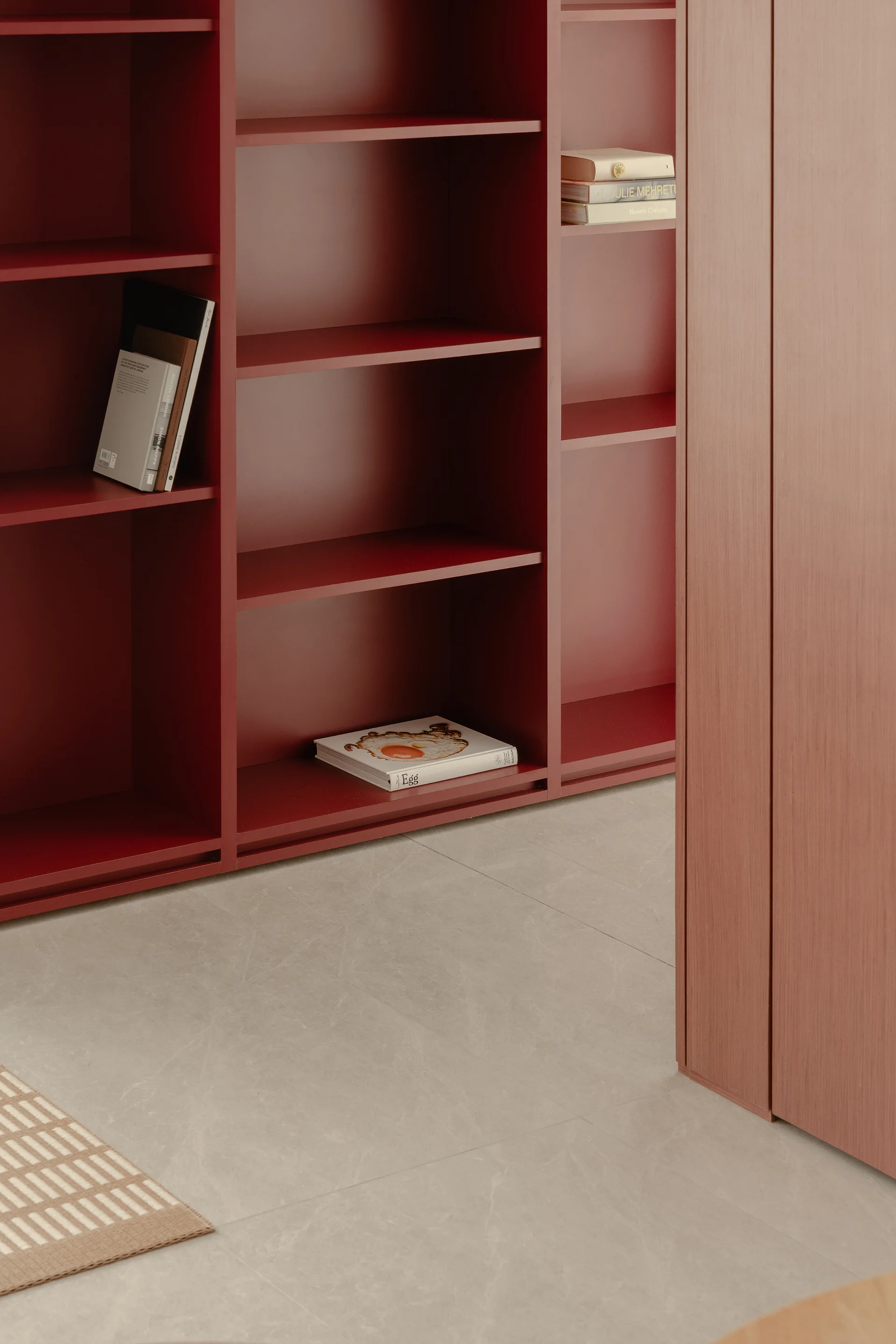 A matte deep red built-in bookshelf features minimalist open shelves alongside a warm wood closet door on a light grey tiled floor.