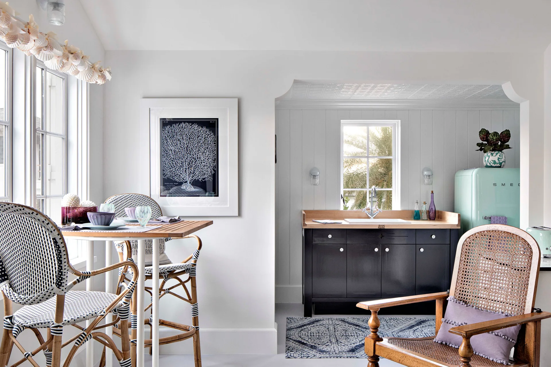 A bright coastal dining nook and kitchenette featuring a light blue retro refrigerator, dark navy cabinets, and woven chairs.