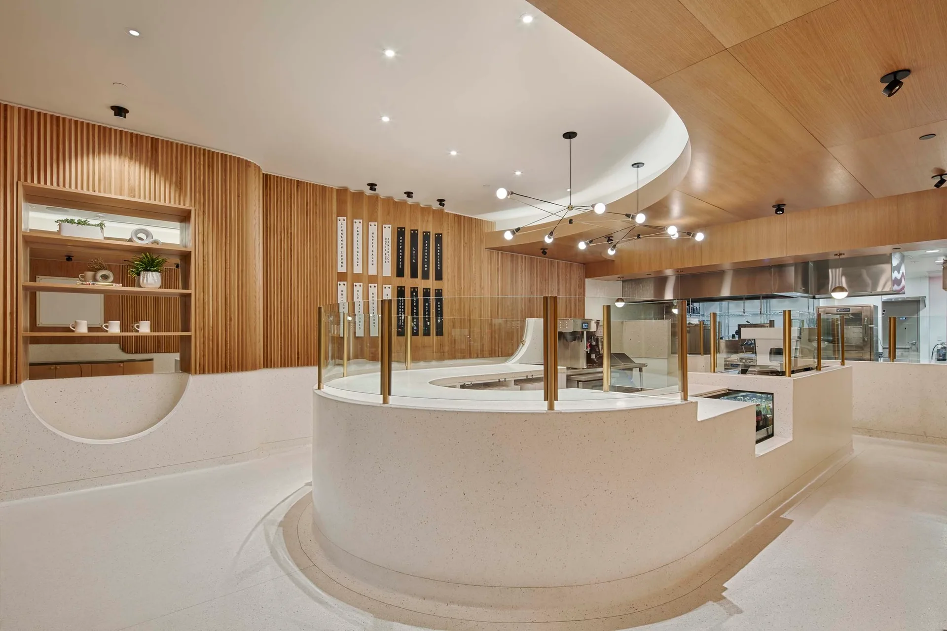 A modern cafe interior featuring a curved speckled counter, a vertical wood slat wall, and a branching chandelier under a recessed ceiling cutout.