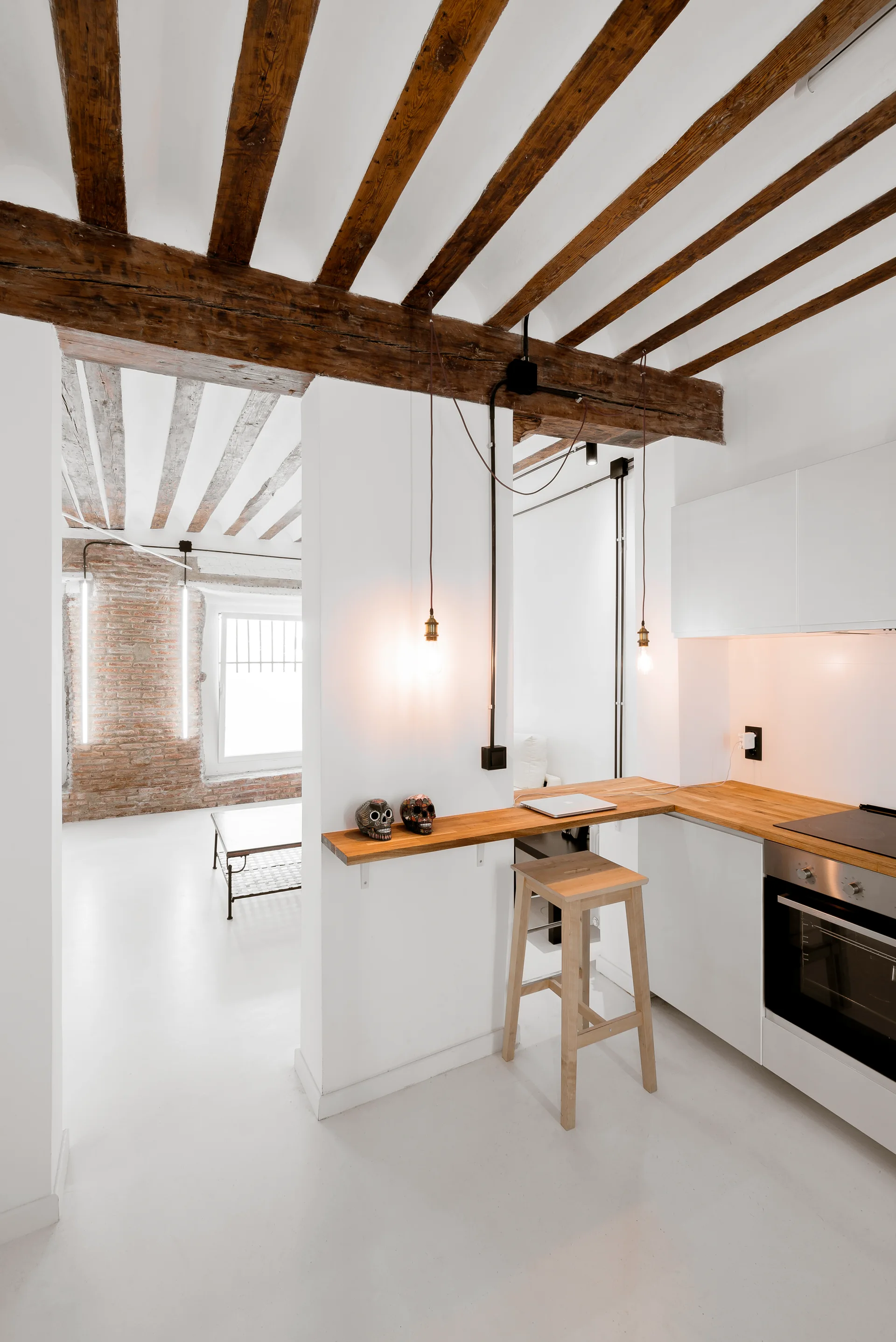 A modern white kitchen features a wooden breakfast bar, minimalist white cabinets, exposed ceiling beams, and a wooden stool.