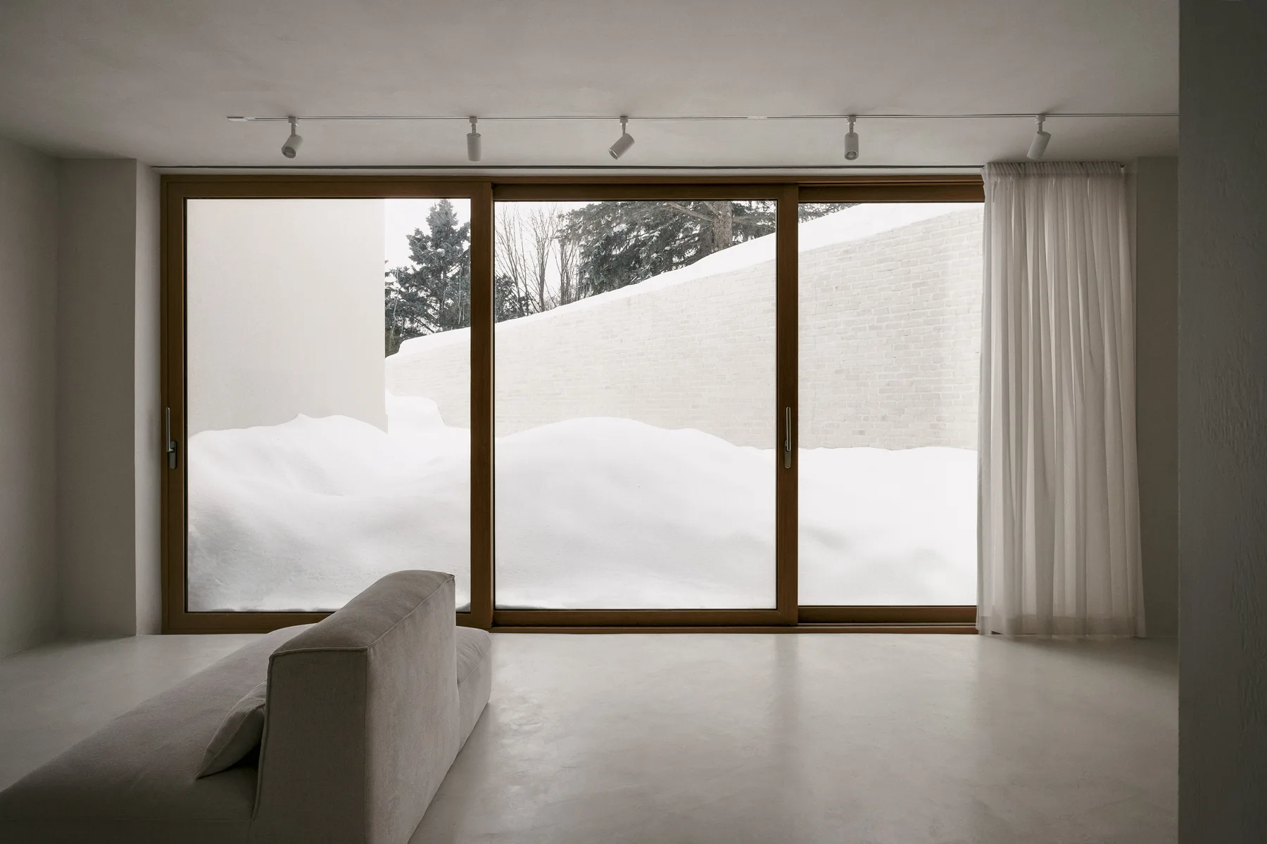 A low-profile beige sofa sits in a minimalist room facing a large wood-framed sliding glass door with a view of a snowy landscape.