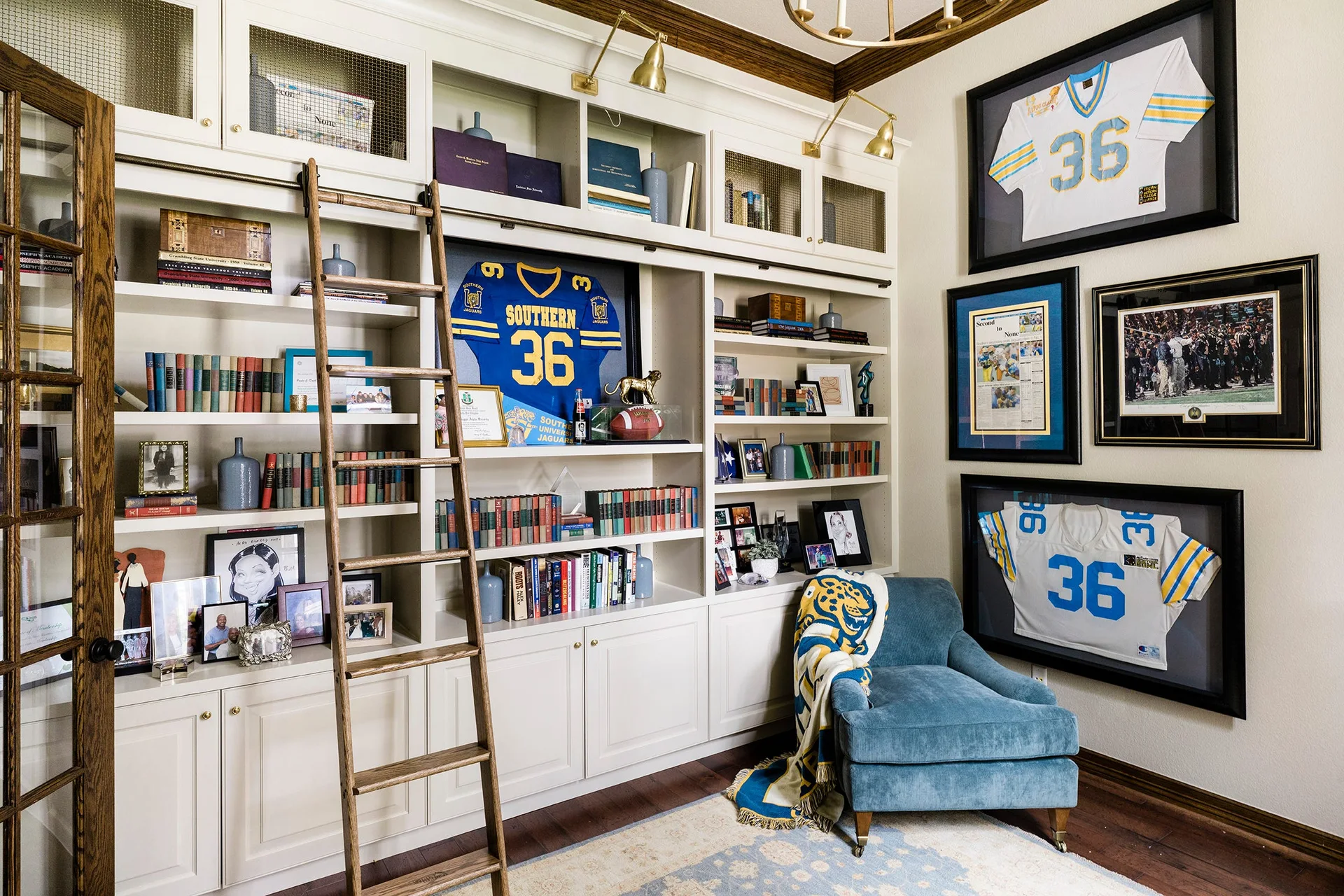 A home library features off-white built-in bookshelves with a wooden rolling ladder, a blue armchair, and framed sports jerseys on the wall.