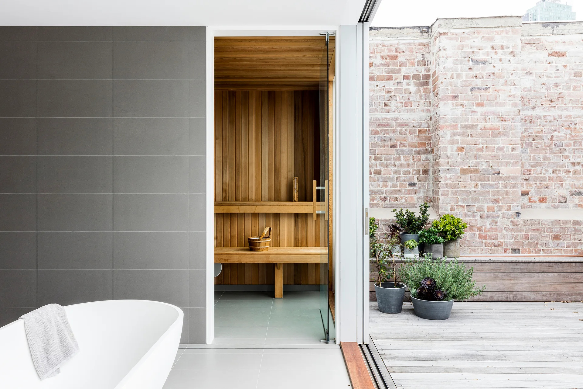 A modern bathroom with a white soaking tub, a wood-lined sauna, and sliding doors opening to an outdoor patio with brick walls and plants.