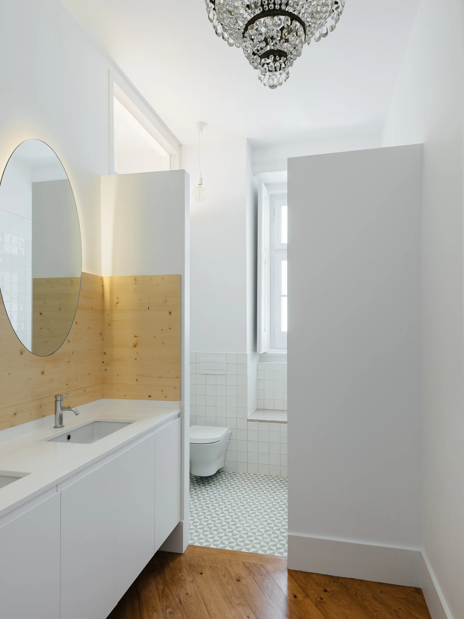 A modern white bathroom featuring a vanity with a light wood backsplash, a round mirror, and a geometric tiled floor leading to a wall-mounted toilet.