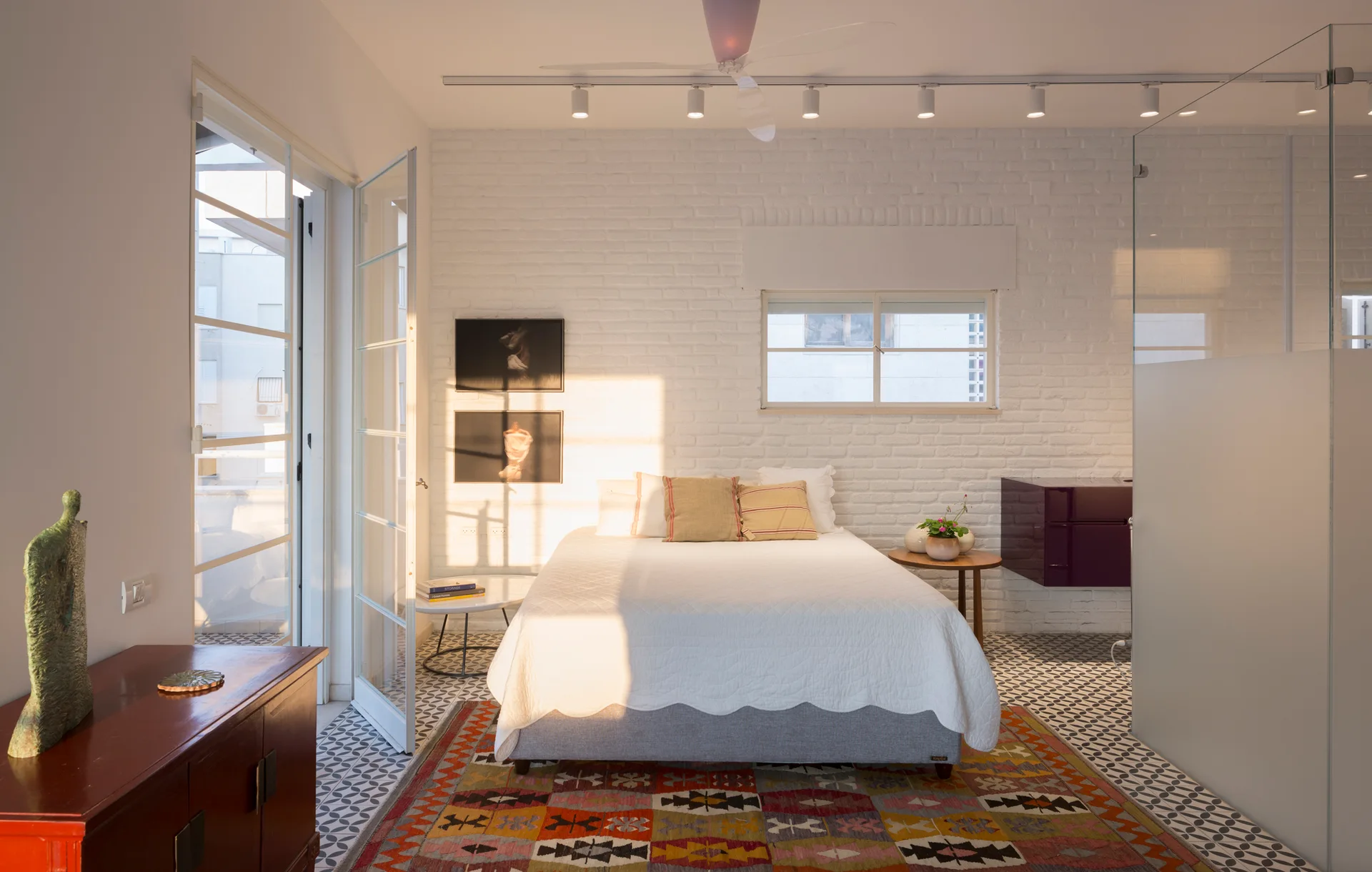 A bedroom features a bed with white linens on a colorful rug, patterned tile flooring, and white painted brick walls with track lighting.