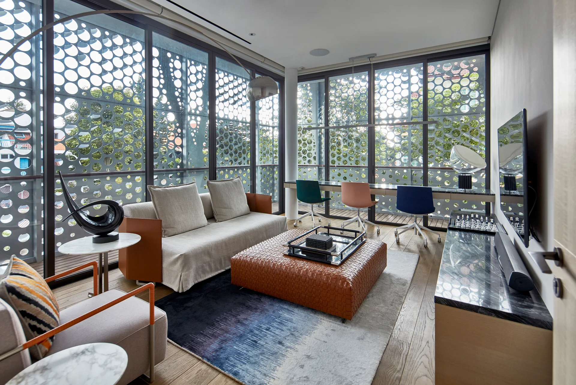 A contemporary living room with a gray sofa, woven leather ottoman, and a home office desk set against large windows with a patterned cutout screen.