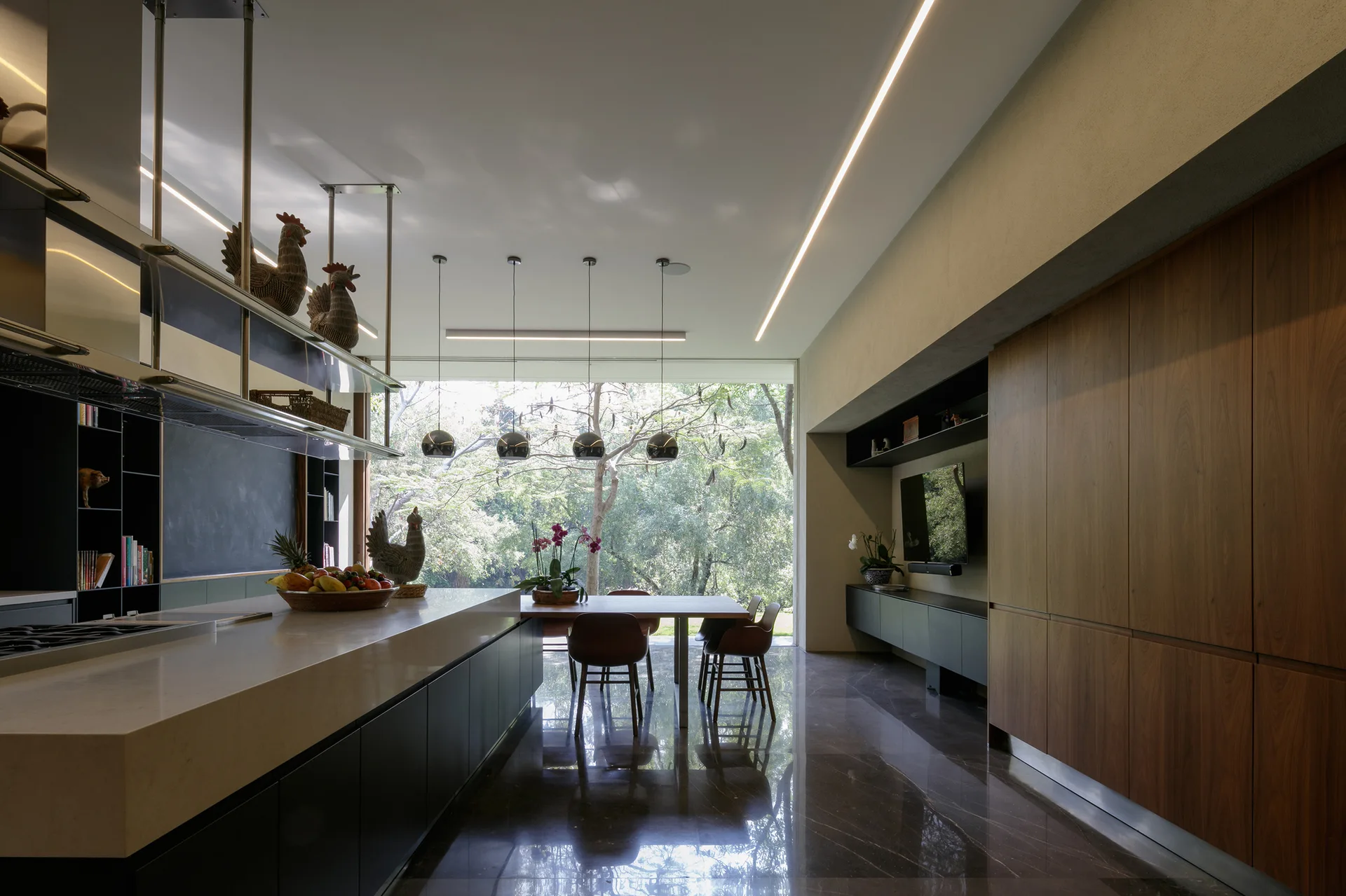 A modern open kitchen and dining area featuring a long stone island, integrated wood cabinetry, and a large window looking out onto trees.