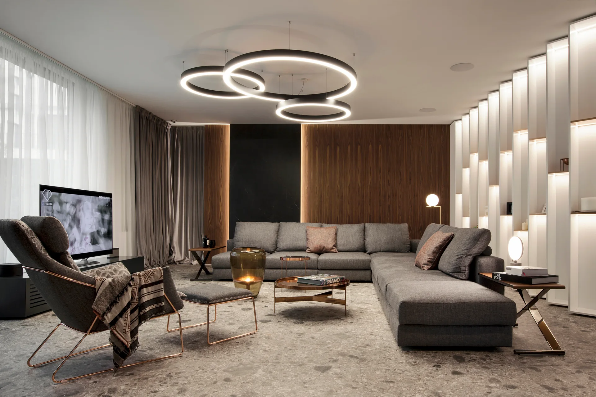 A contemporary living room features a gray sectional sofa, large ring pendant lights, a wood-paneled accent wall, and stone-look tile flooring.