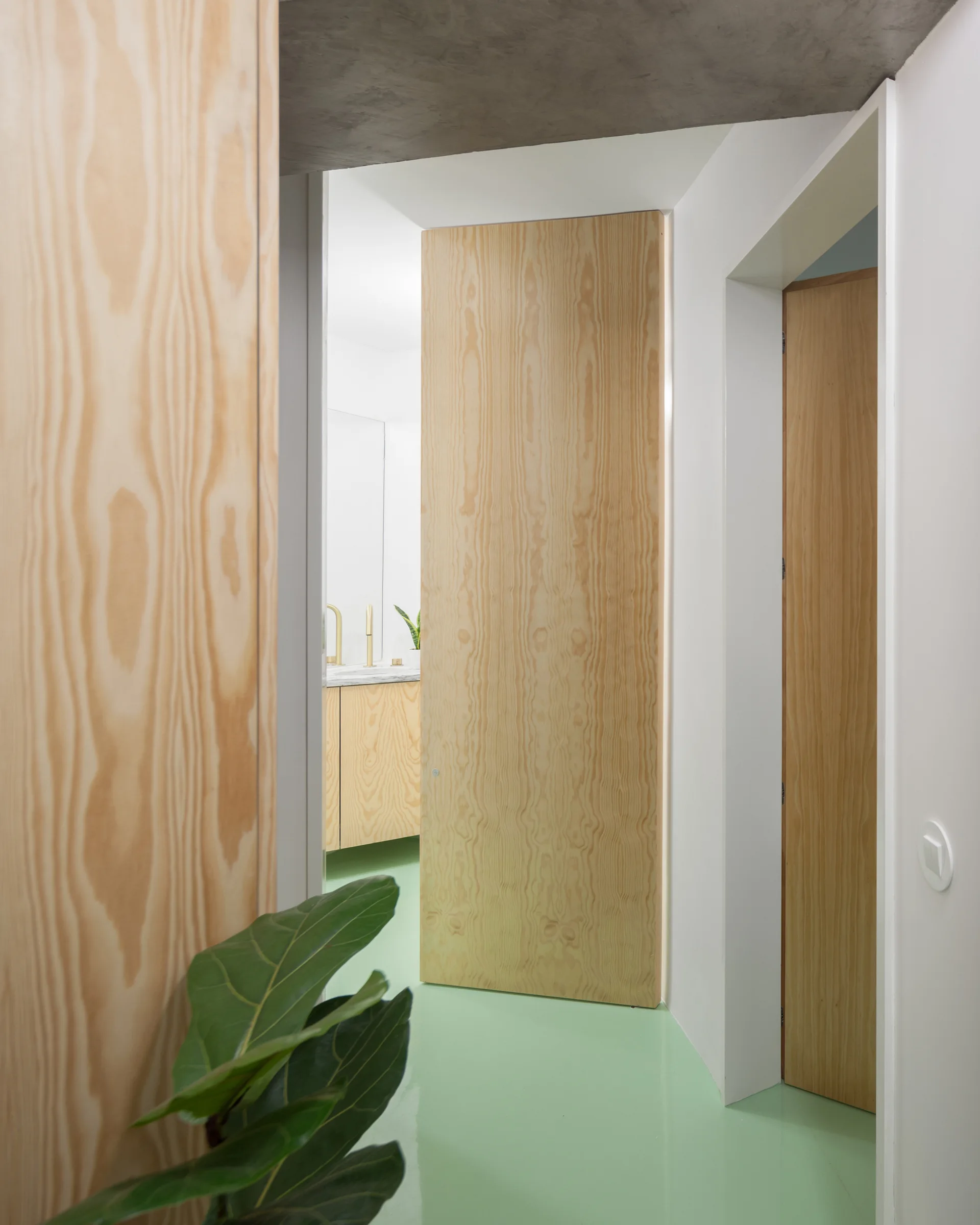 A minimalist hallway features high-gloss mint green floors, light wood veneer walls with flush doors, and a glimpse of a bathroom vanity in the background.
