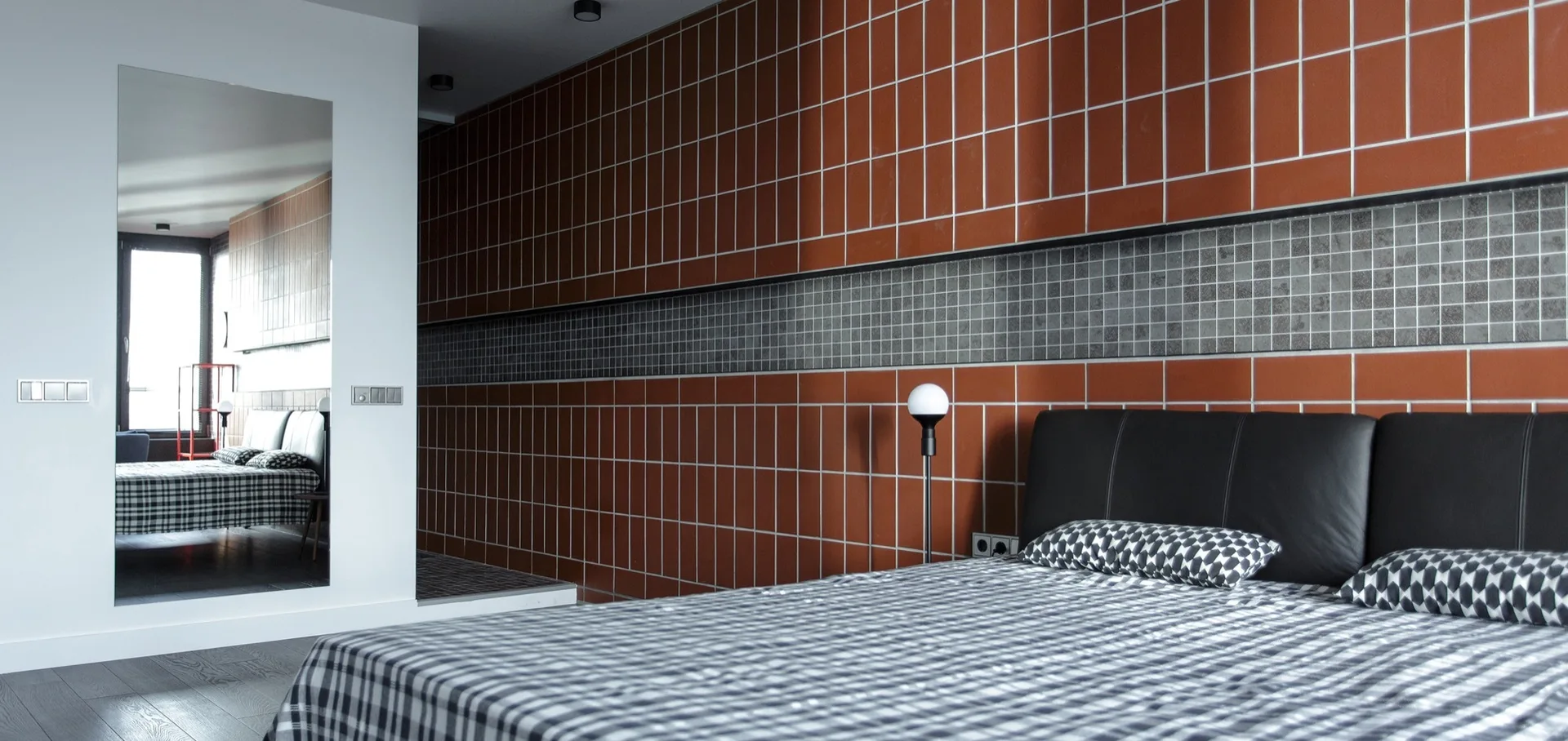 A modern bedroom features a red and grey tiled accent wall, a bed with a black headboard and plaid bedding, and a tall rectangular wall mirror.