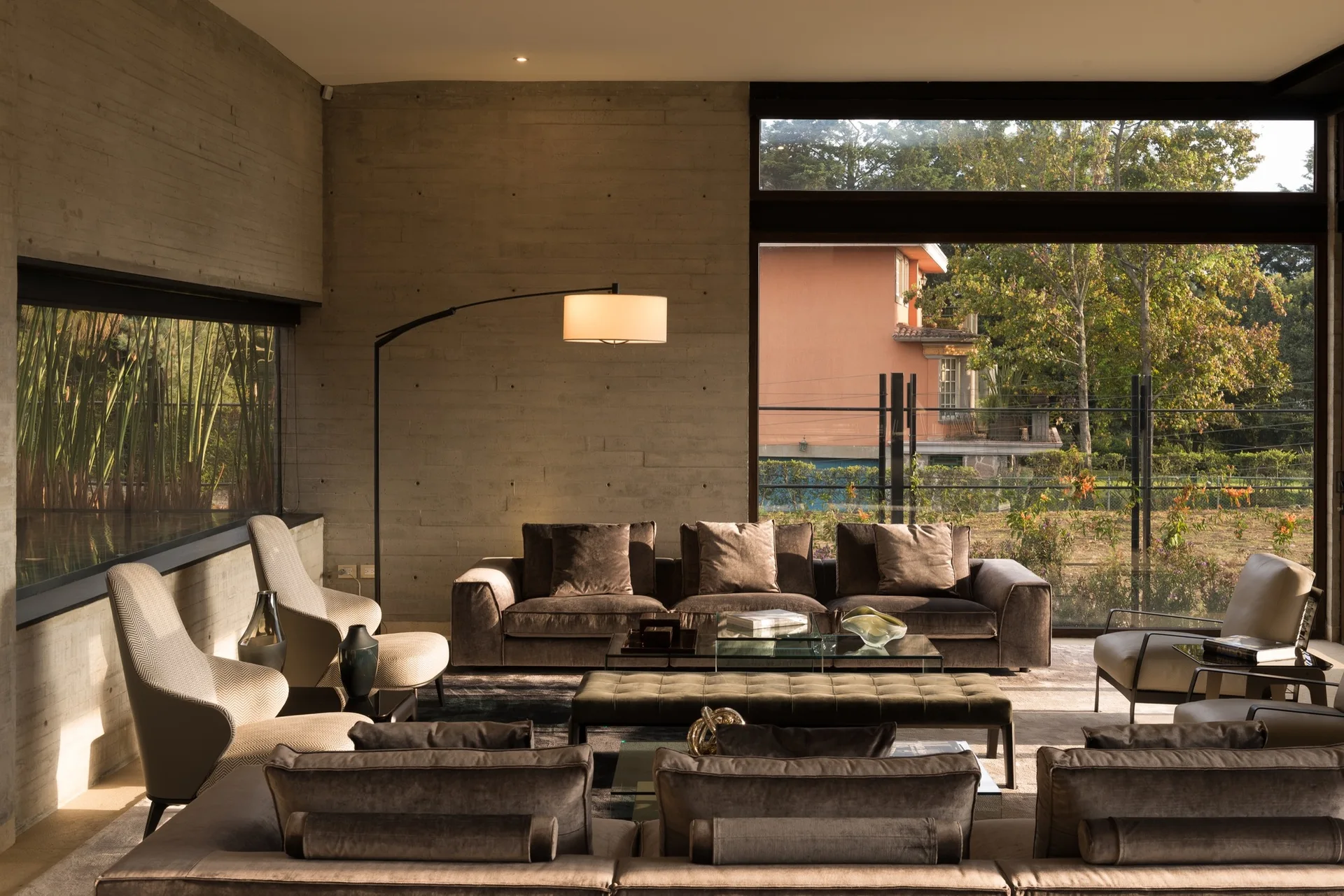 A modern living room features textured concrete walls, deep brown velvet sofas, a glass coffee table, and large windows looking out to a garden.