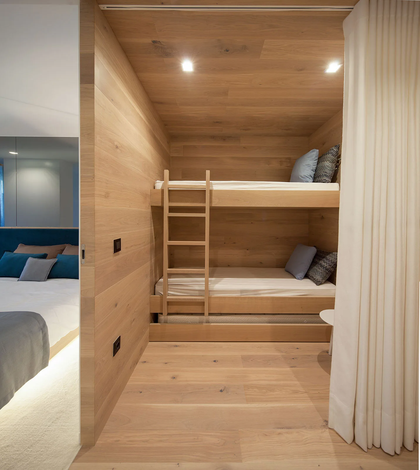 Built-in wooden bunk beds are nestled within a fully wood-paneled alcove next to a bedroom, featuring a ladder and a white privacy curtain.