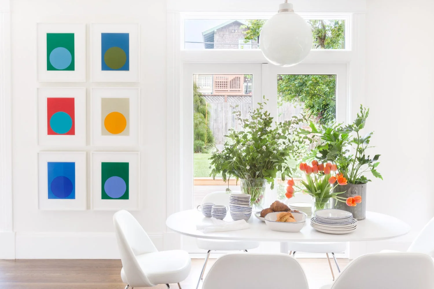 A bright white dining room featuring an oval table, modern white chairs, a globe pendant light, and a grid of six colorful geometric art prints.