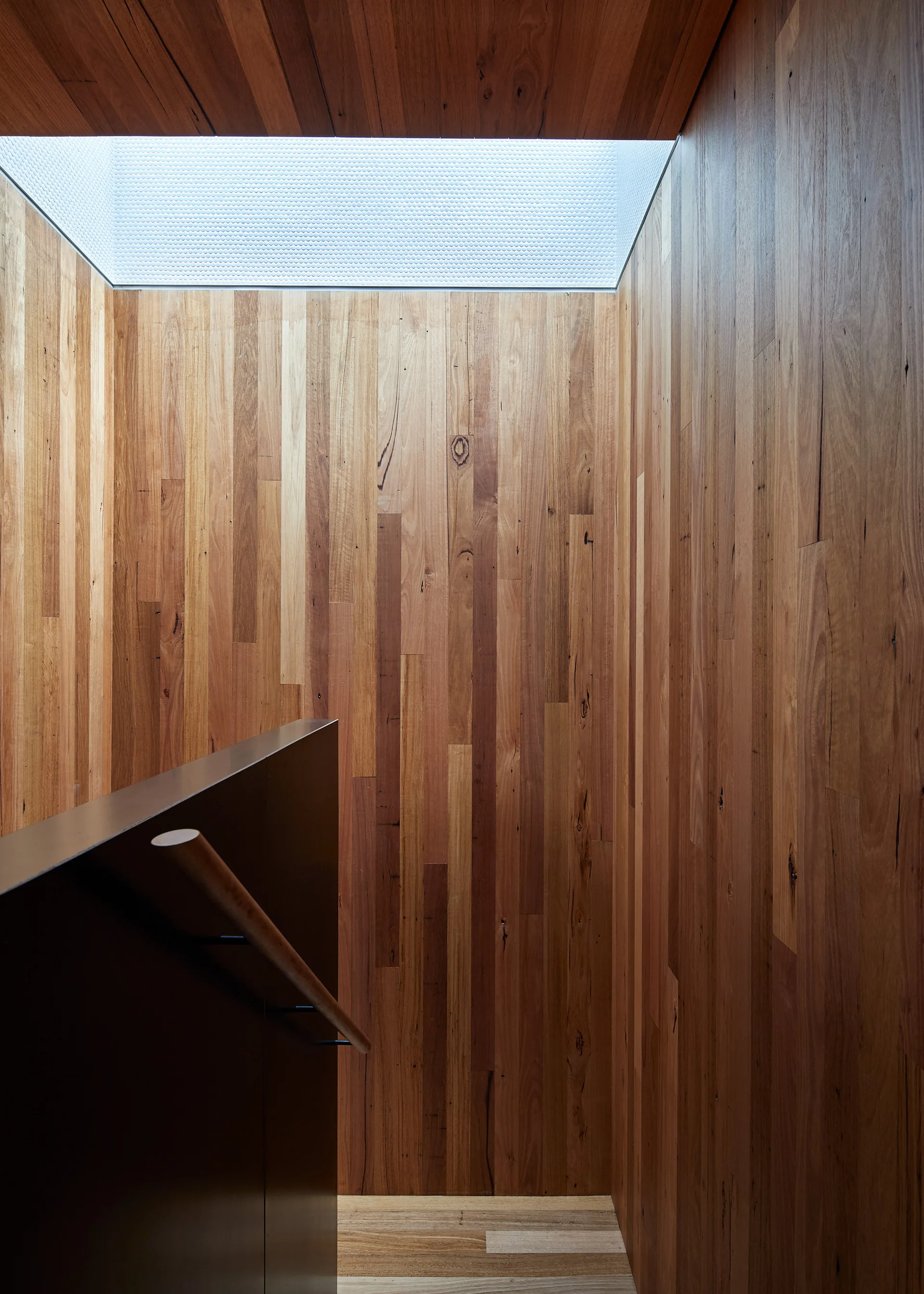 A minimalist stairwell features warm vertical wood paneled walls, a dark railing with a round handrail, and a rectangular skylight above.