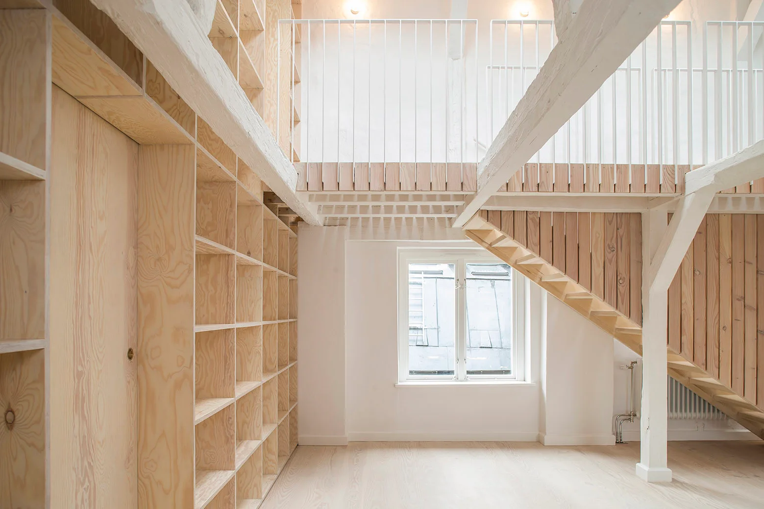 A bright two-level interior featuring a large wall of light wood plywood shelving, a matching wooden staircase, and white ceiling beams.