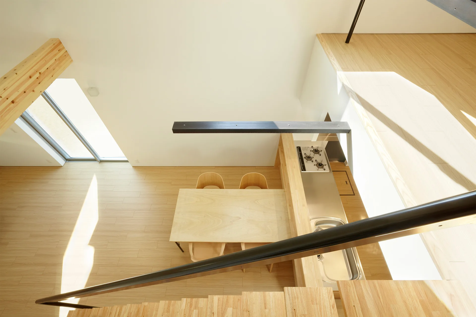 A high-angle view looking down at a minimalist kitchen with a stainless steel counter, light wood dining table, and stairs in the foreground.