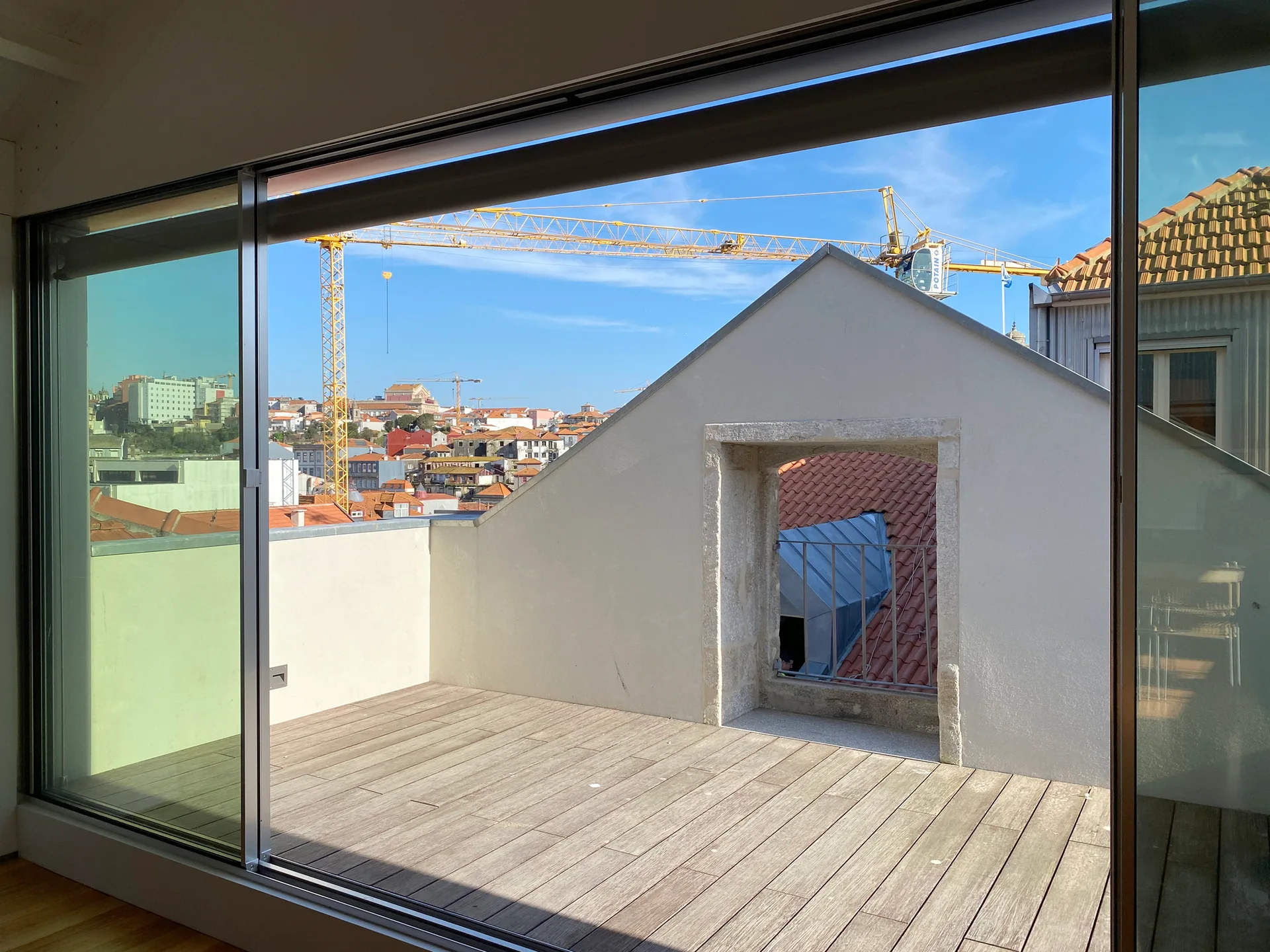 A view through a large glass sliding door onto a wooden plank terrace with a white stucco wall and a distant city skyline.